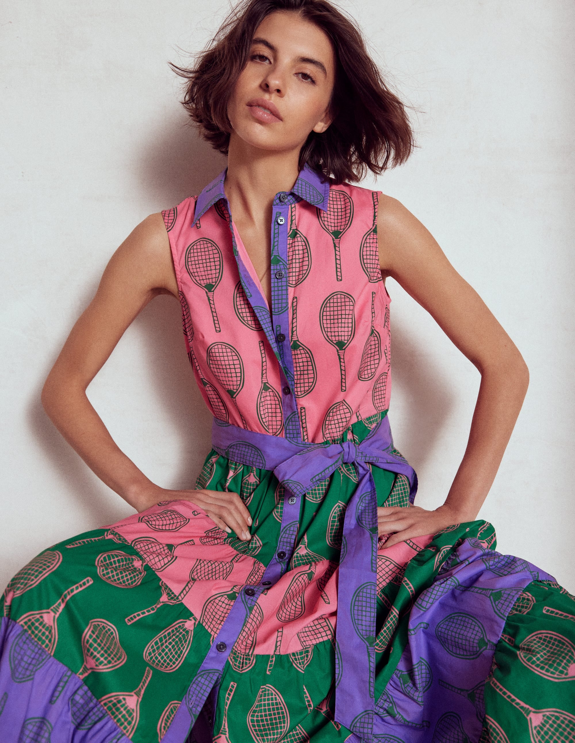 A woman wearing a sleeveless, pink shirt dress with tennis racket print, purple collar, and a purple belt, standing against a plain background.