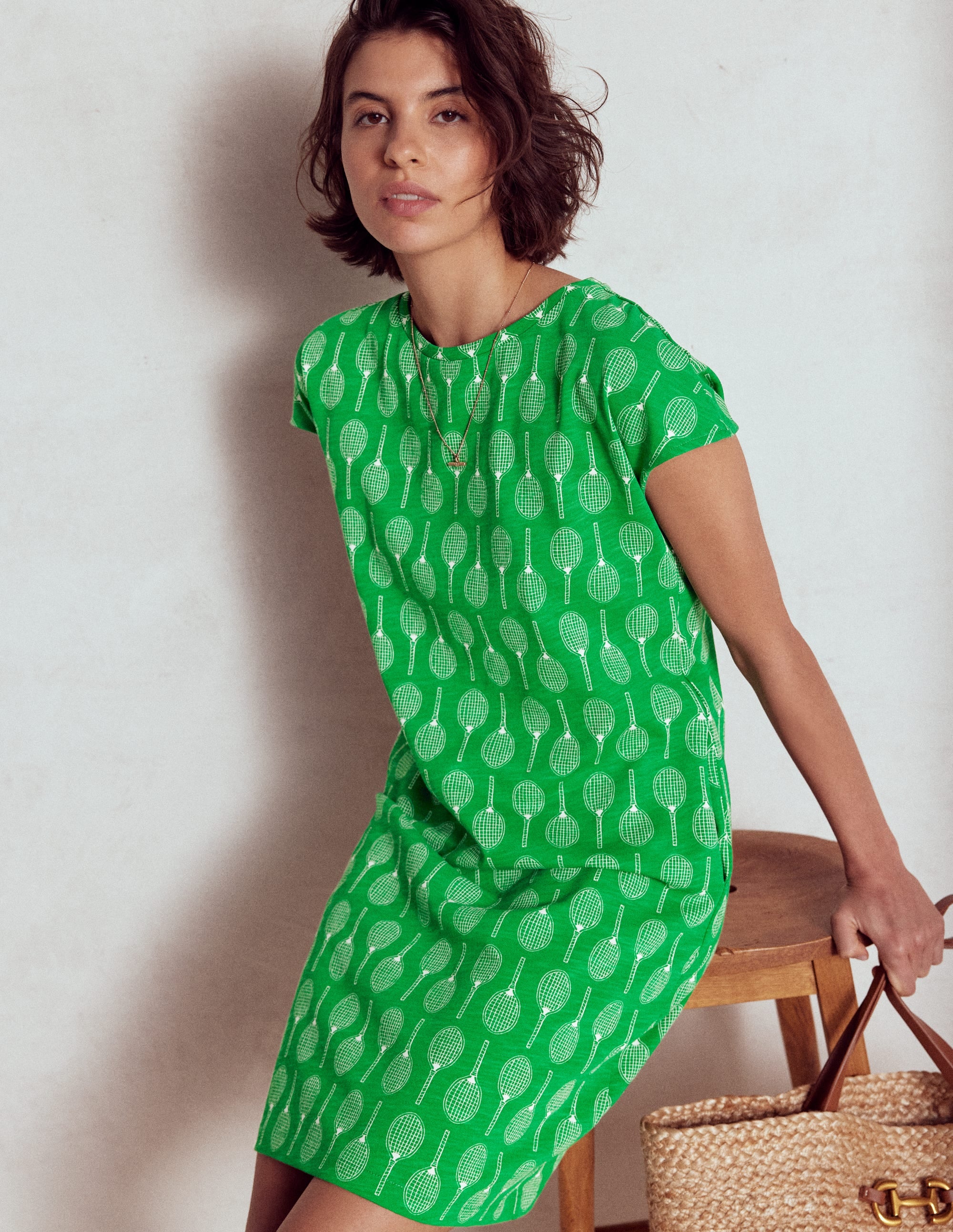 A woman wearing a green T-shirt dress with tennis racket patterns, sitting on a wooden chair, holding a woven bag.