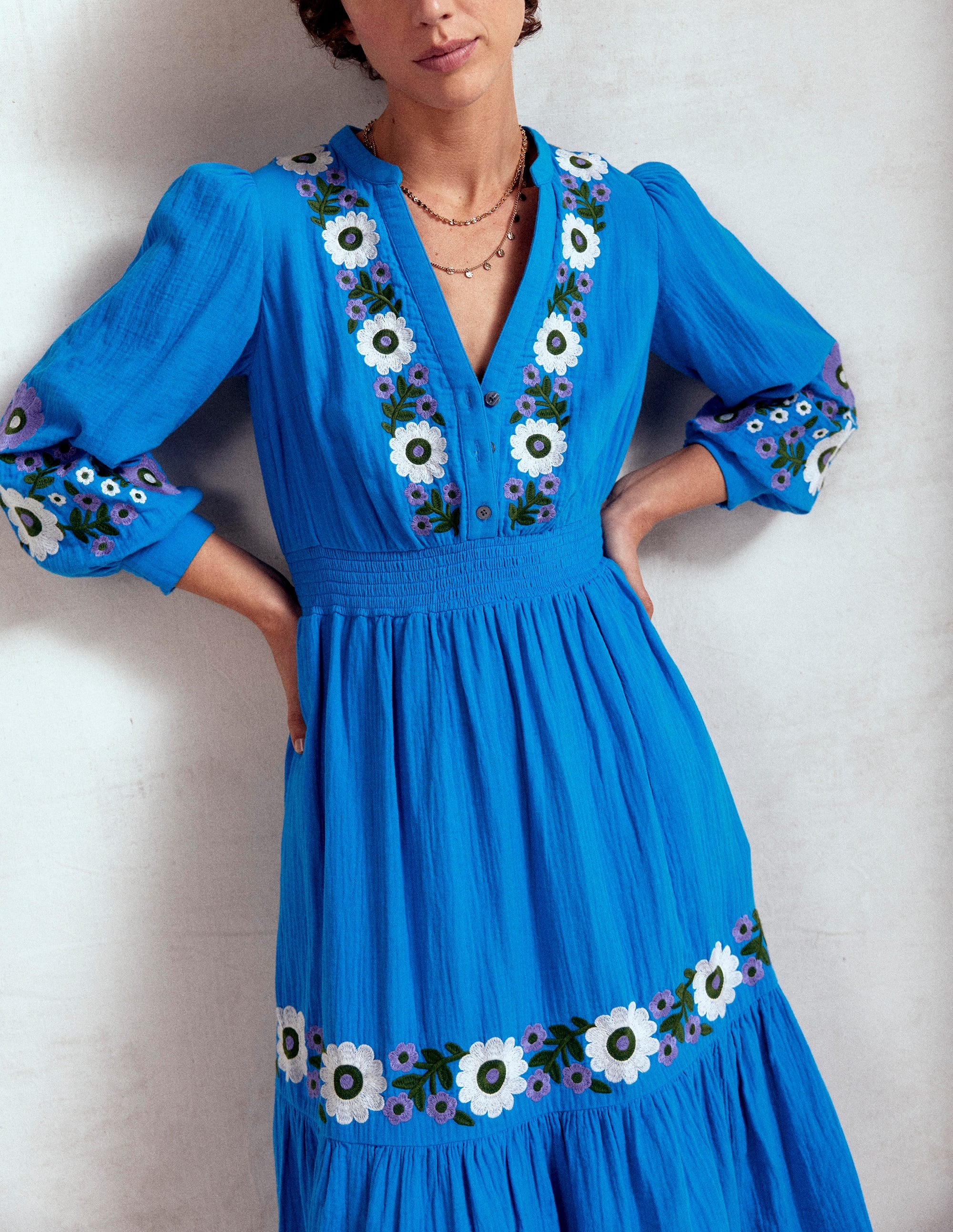 A woman wearing a bright blue embroidered midi dress with floral patterns on the front, sleeves, and hem, with a V-neckline and long sleeves.
