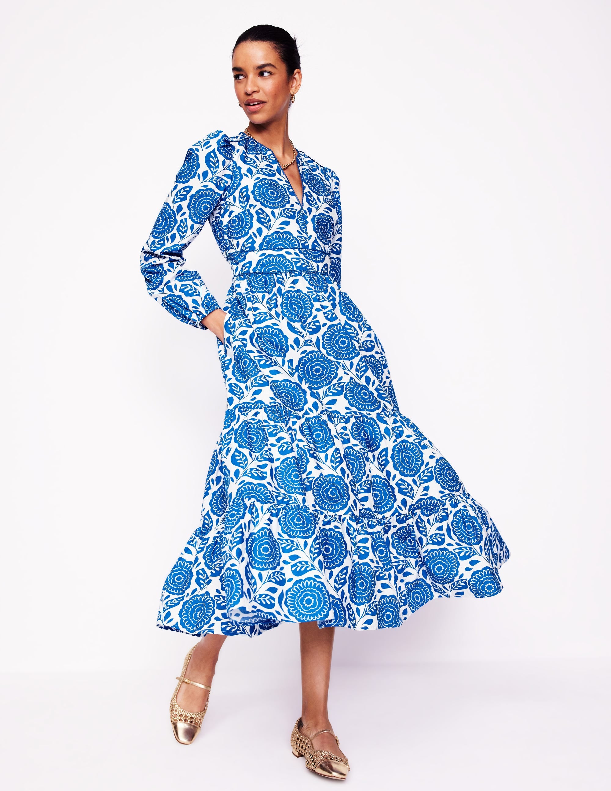 A woman wearing a blue floral midi dress with long sleeves, V-neckline, and side pockets, standing against a white background.