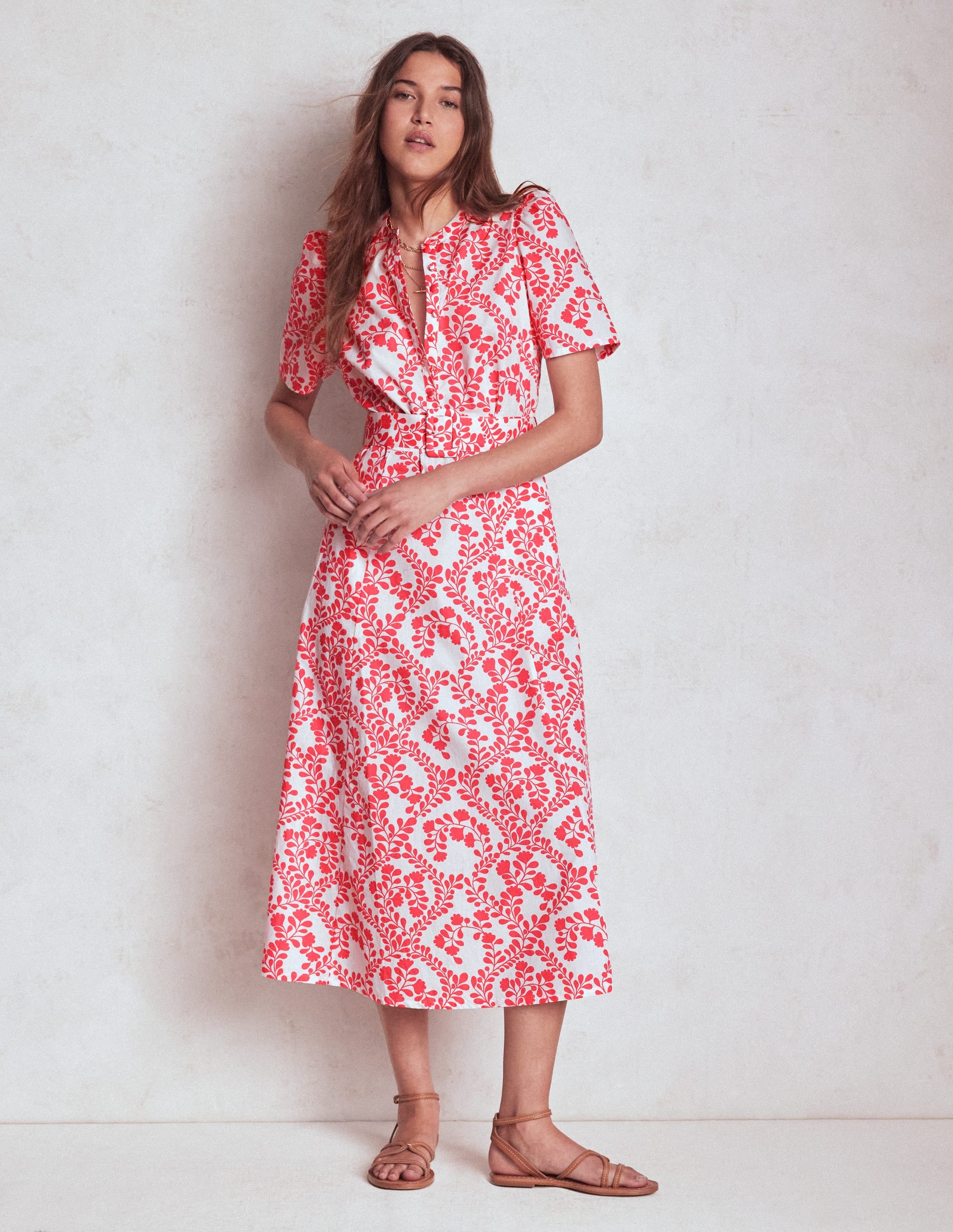 A woman wearing a belted dress with puff sleeves and a vine pattern in ivory and coral, standing against a plain background.