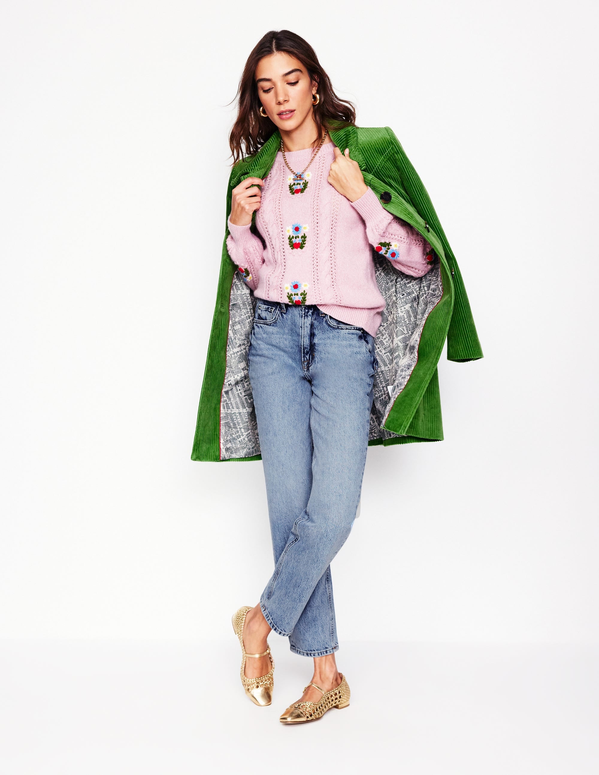 A woman wearing light vintage blue high-rise straight-leg jeans, a pink embroidered sweater, and a green coat, standing against a white background.