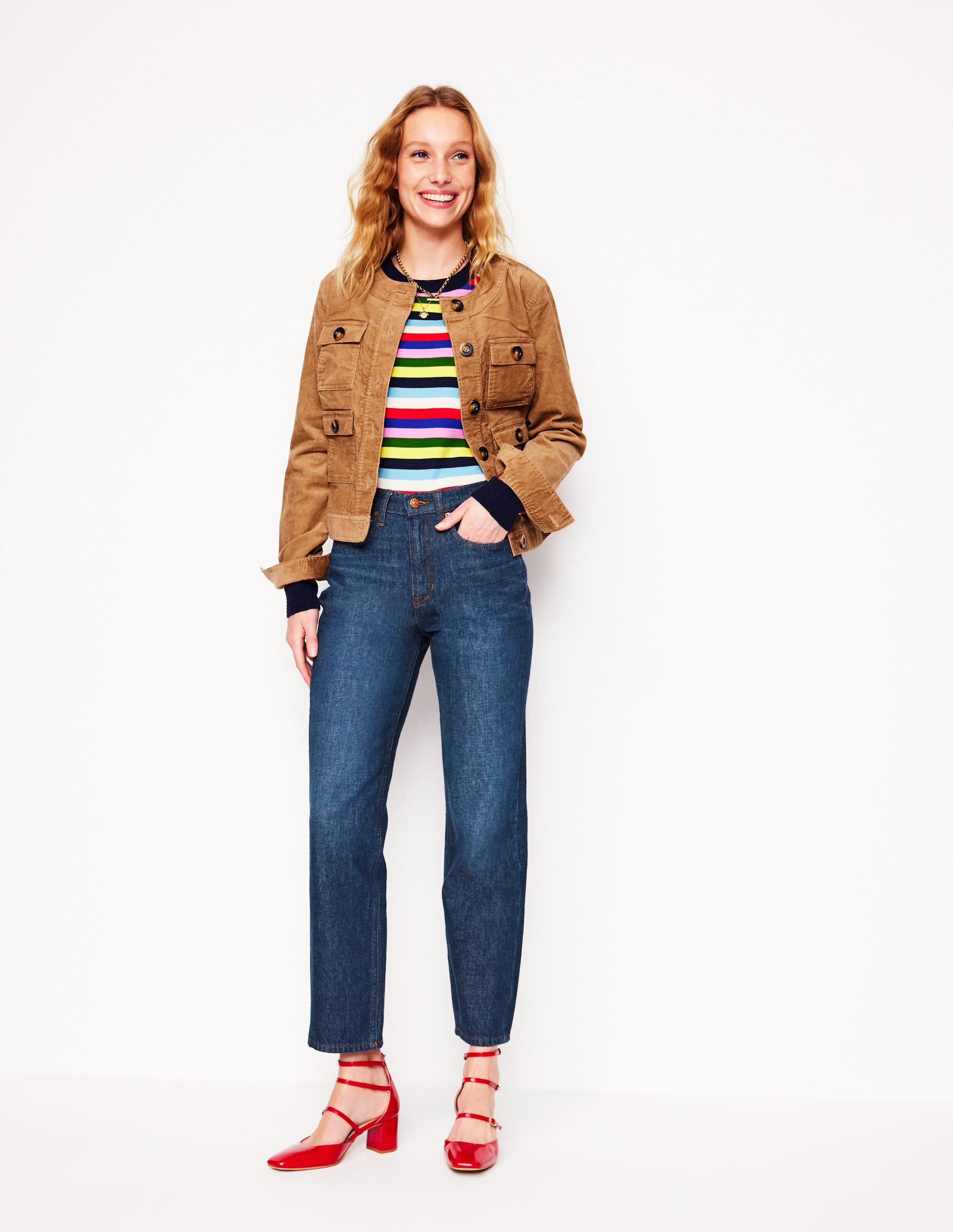 A woman wearing high-rise indigo rinse straight-leg jeans, a colorful striped top, a tan jacket, and red heels.