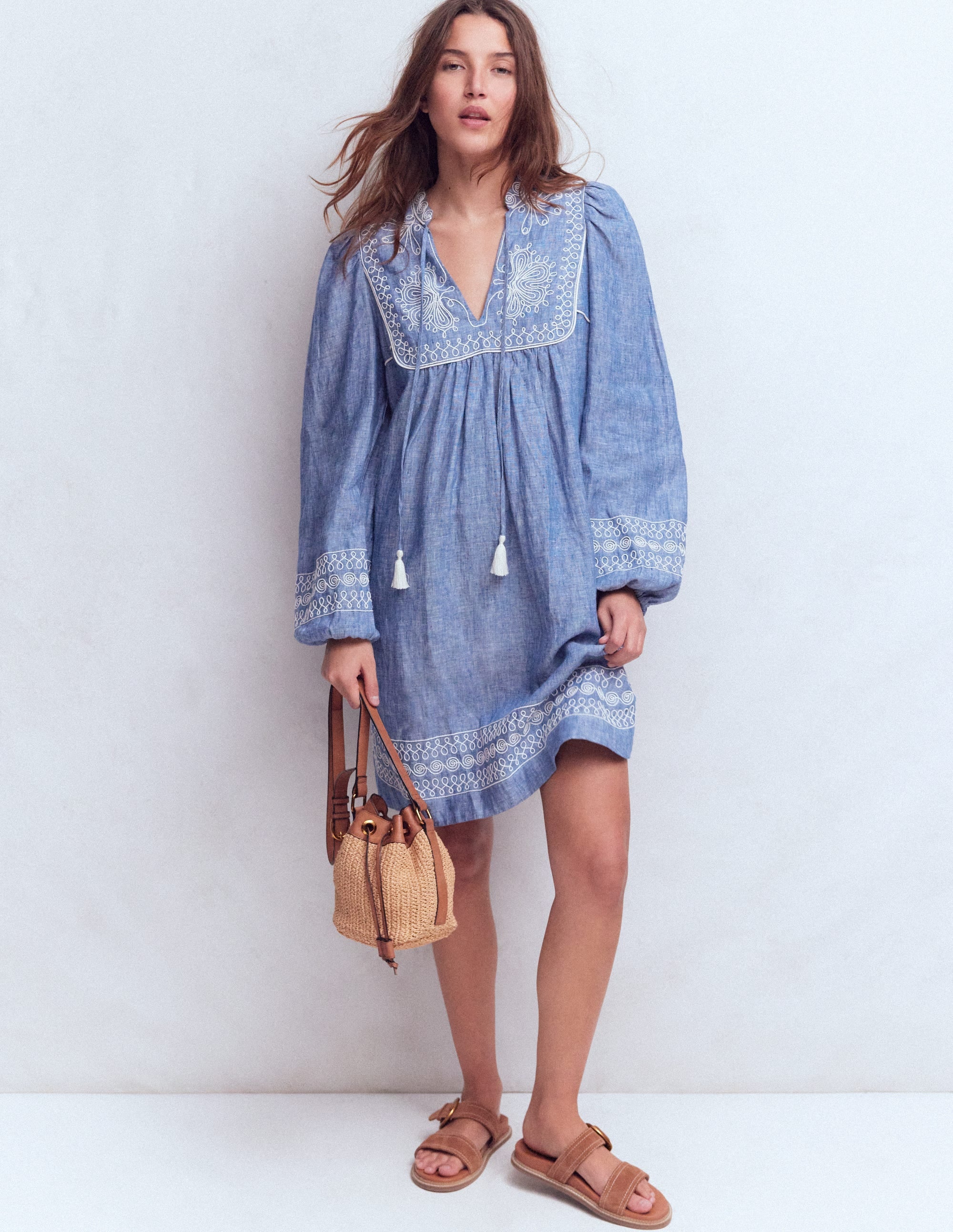 A woman wearing a blue chambray linen dress with white embroidery on the yoke and cuffs, long sleeves, and a relaxed fit, paired with tan sandals and a woven bag.