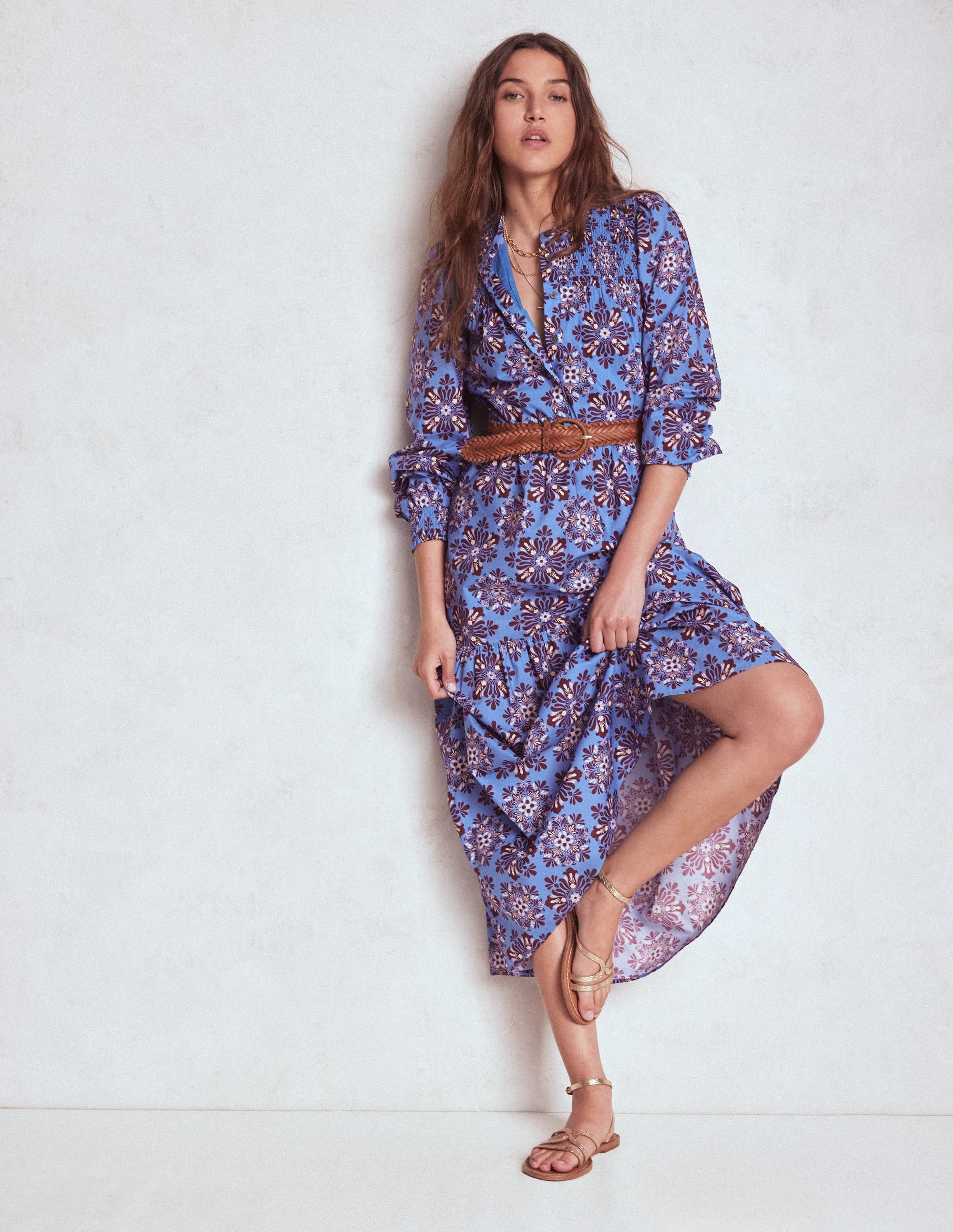 A woman wearing a soft blue floral mosaic midi dress with a fit and flare silhouette, long sleeves, and a belt, standing against a plain background.