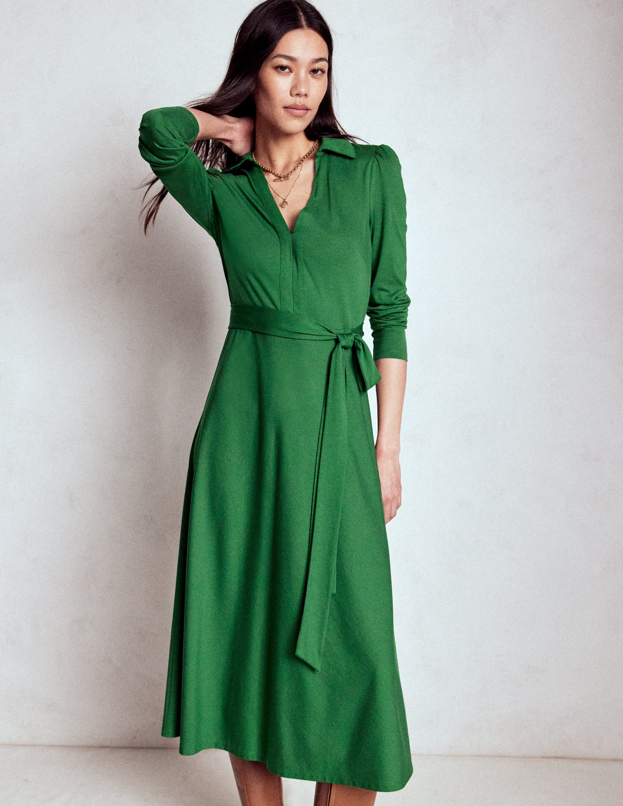 A woman wearing a pine green maxi dress with a collar, long sleeves, and a self-fabric belt, standing against a plain background.