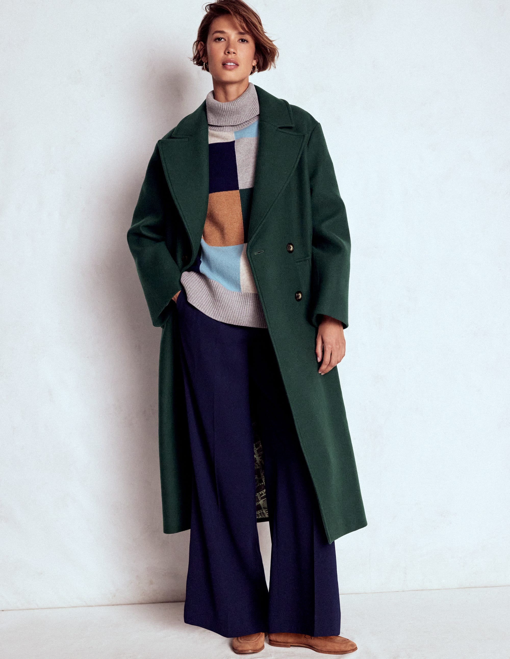 A woman wearing a long emerald green wool coat with a large collar and cocoon shape, paired with a multicolored sweater and navy wide-leg pants.
