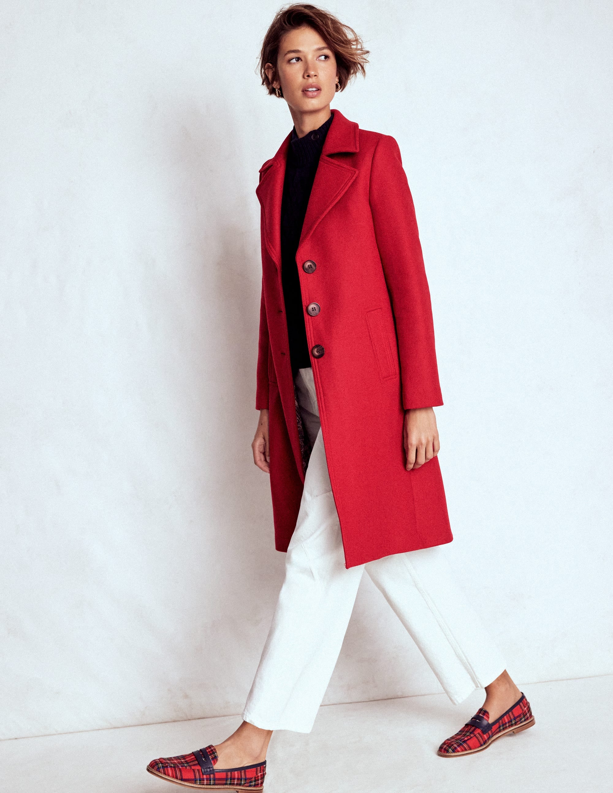 A woman wearing a long, red wool coat with a notched collar, button closure, and welted pockets, paired with white pants and plaid shoes.