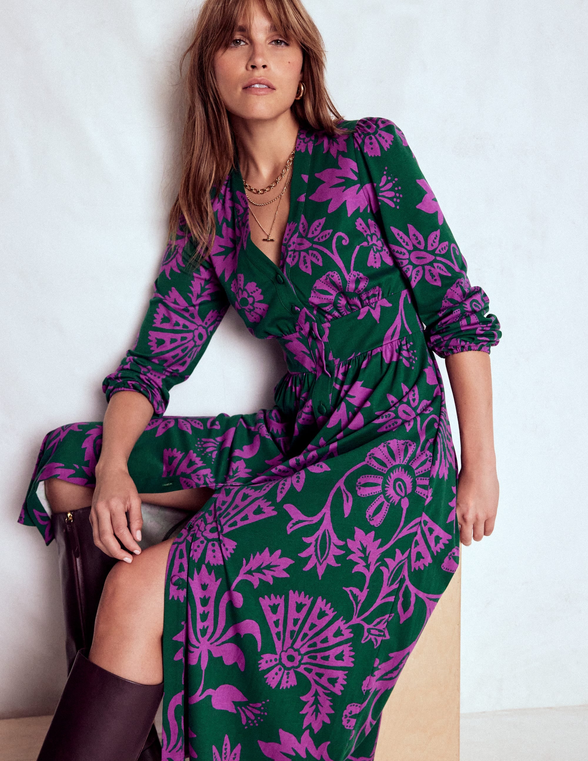 A woman wearing a long sleeve tea dress with a purple vine print on an emerald night background, featuring a fitted waist and flowing skirt, with a lined bodice and side pockets.