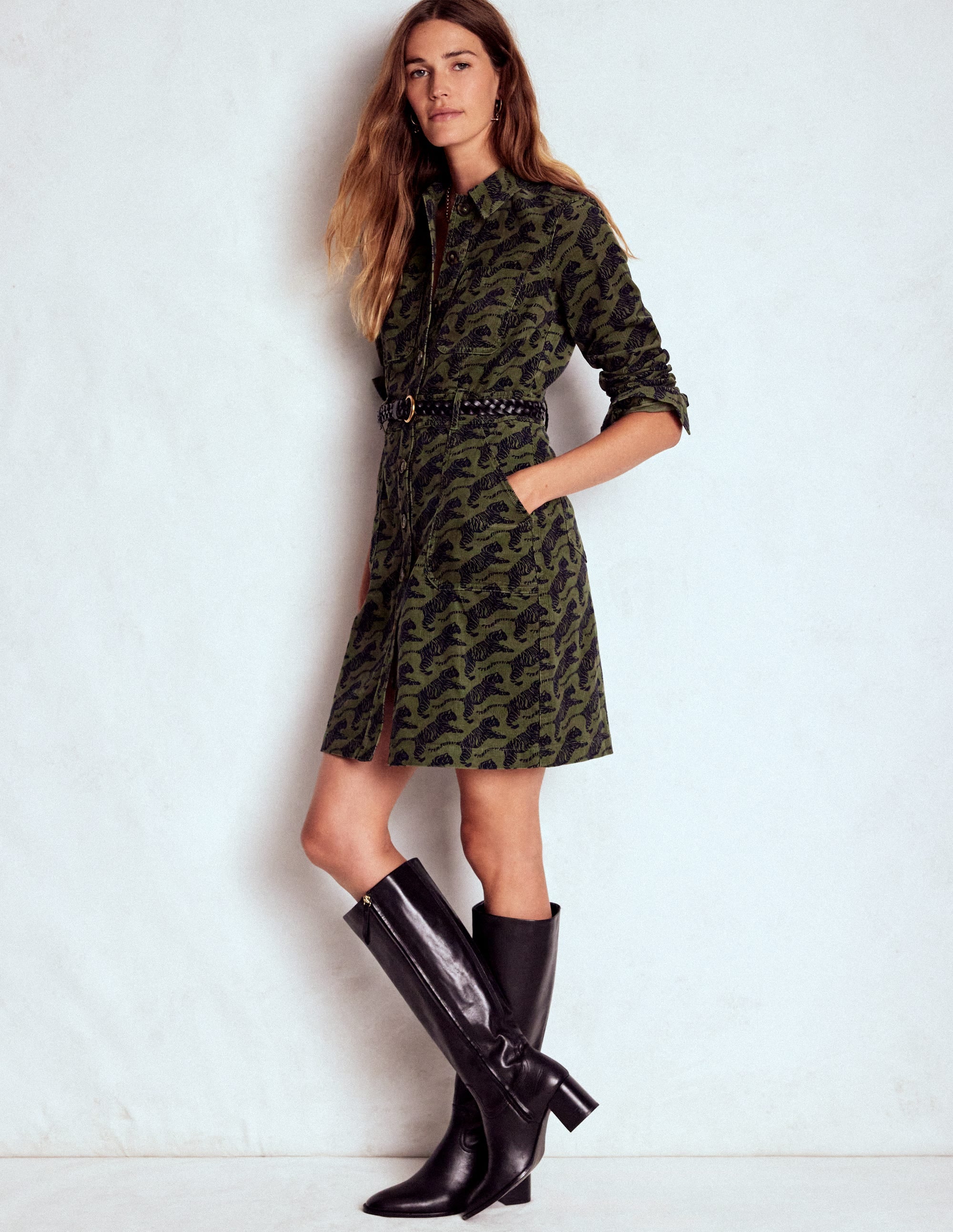 A woman wearing a woodland green short dress with a black tiger pattern, utility details, and a front zipper, paired with black knee-high boots.