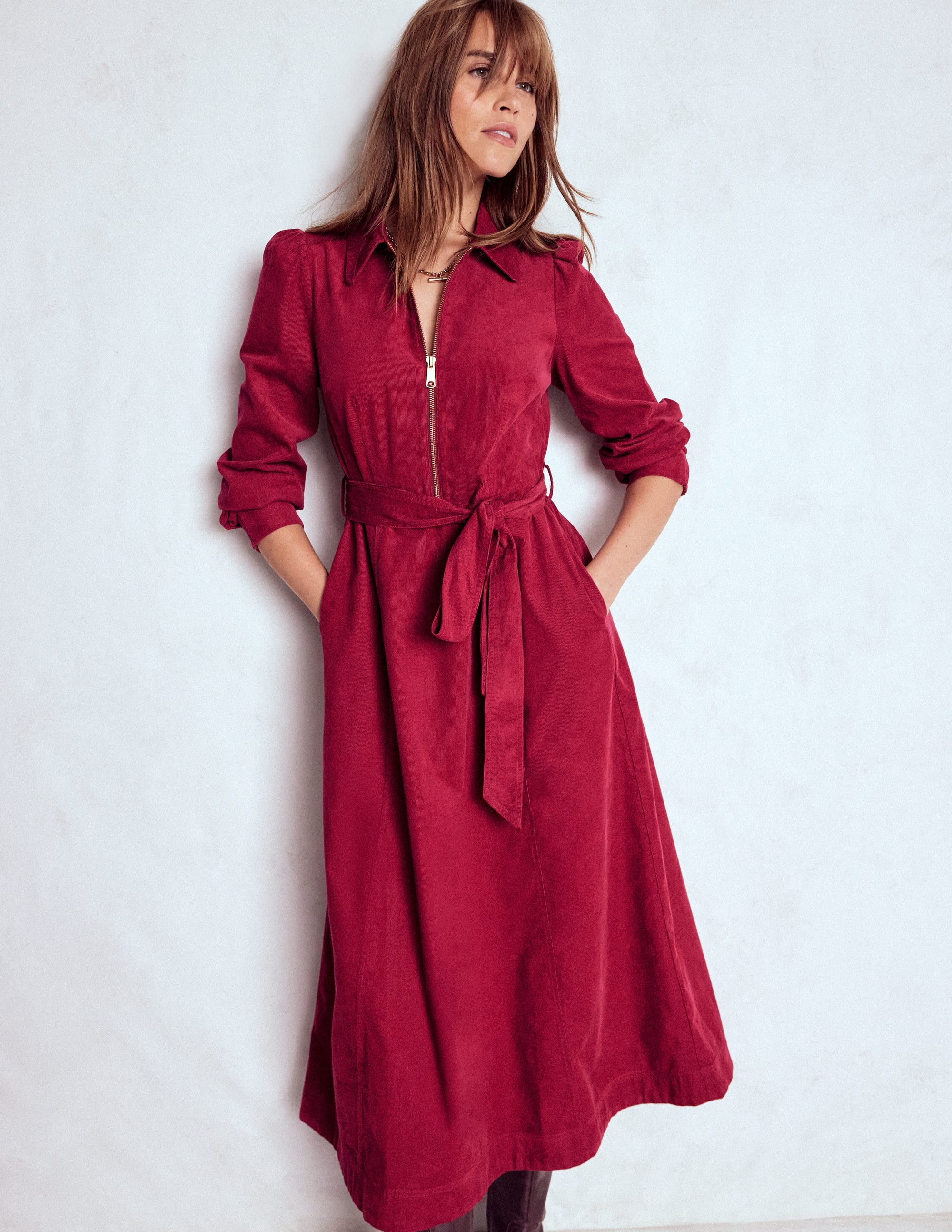 A woman wearing a burgundy red midi dress with a zip-up top, fabric belt, and pockets, standing against a plain background.