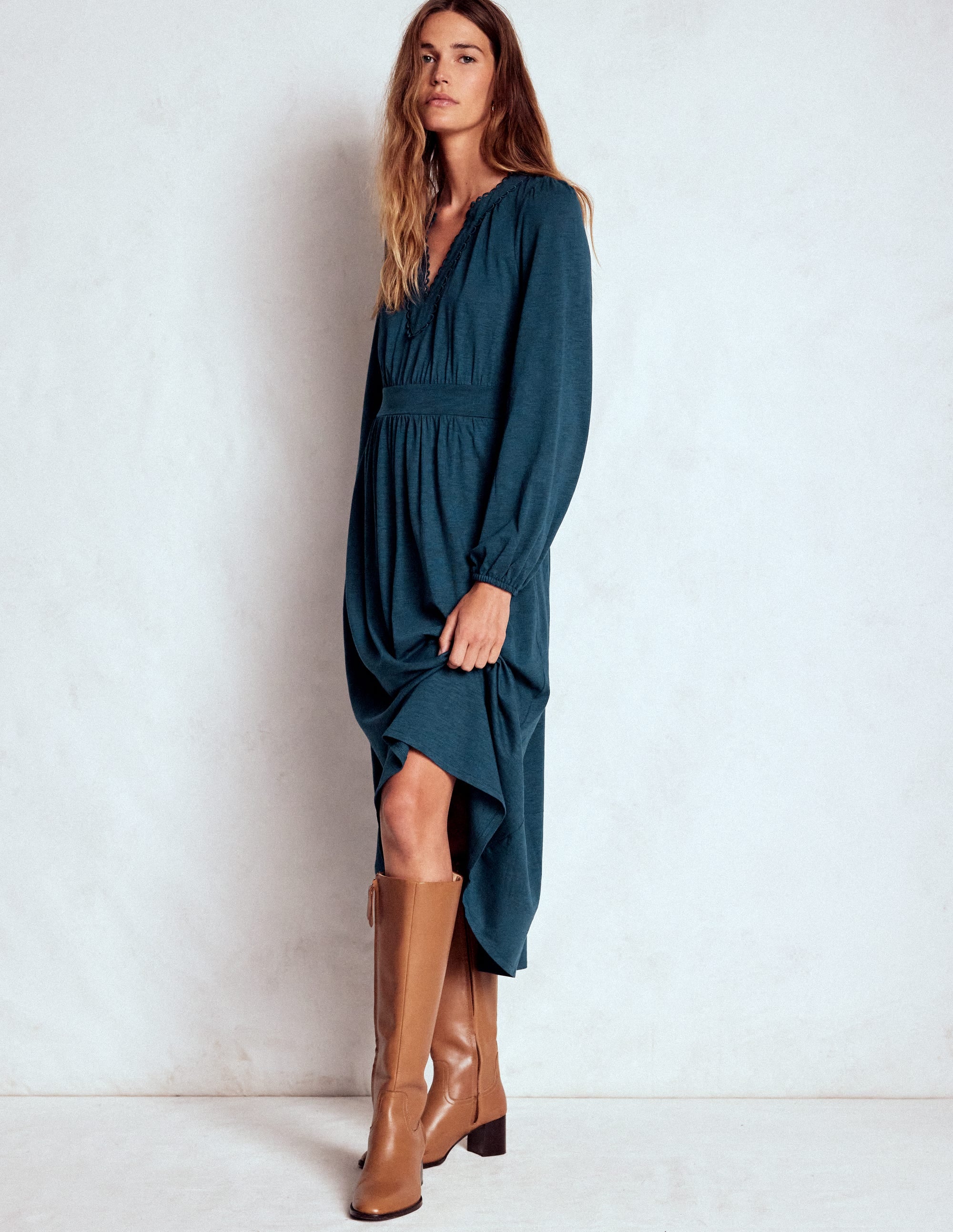 A woman wearing a midnight teal midi dress with blouson sleeves, picot trim, and side seam pockets, paired with tan knee-high boots.