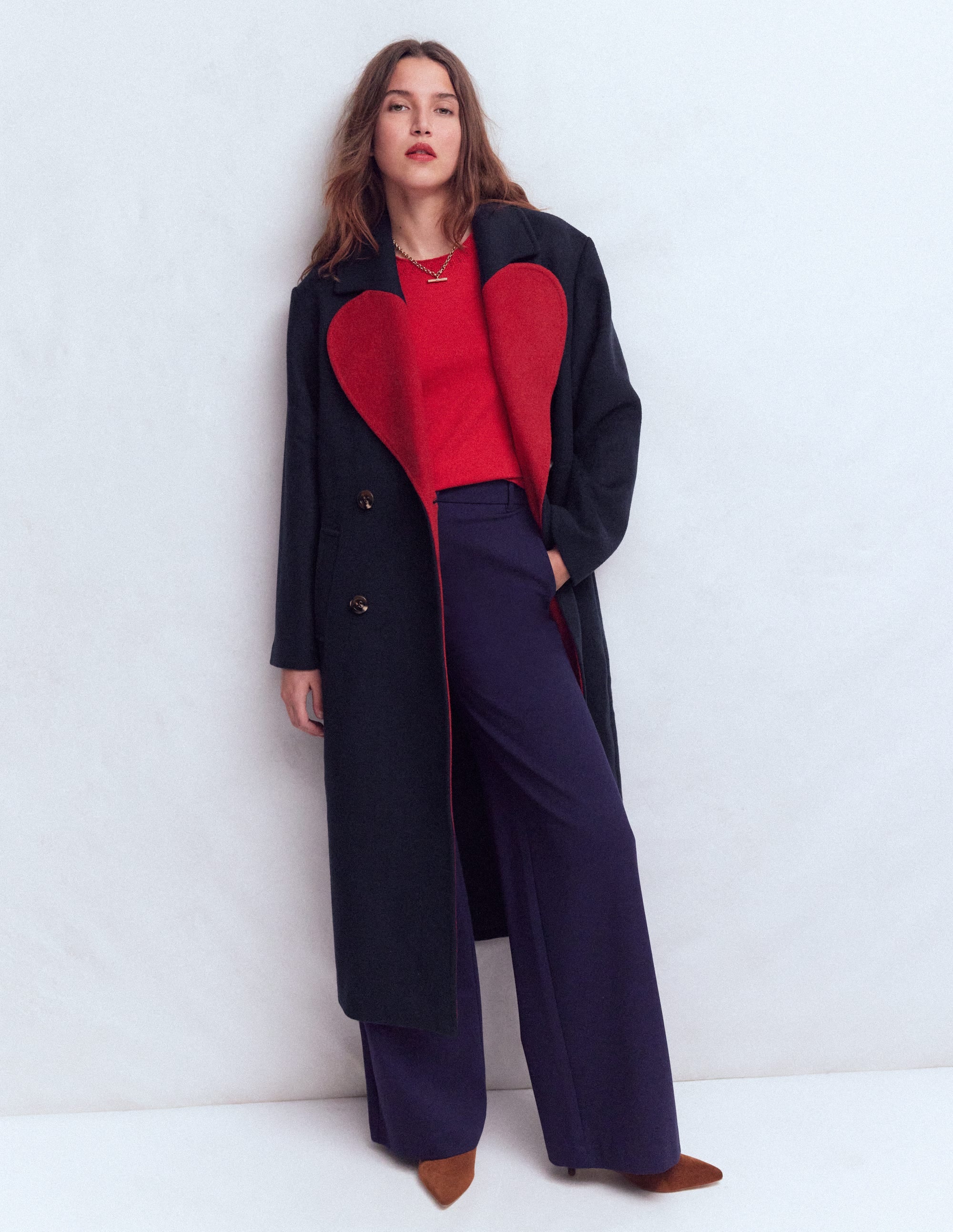 A woman wearing a long navy wool coat with a large red heart on the lapels, double-breasted, with pockets, and an oversized fit.