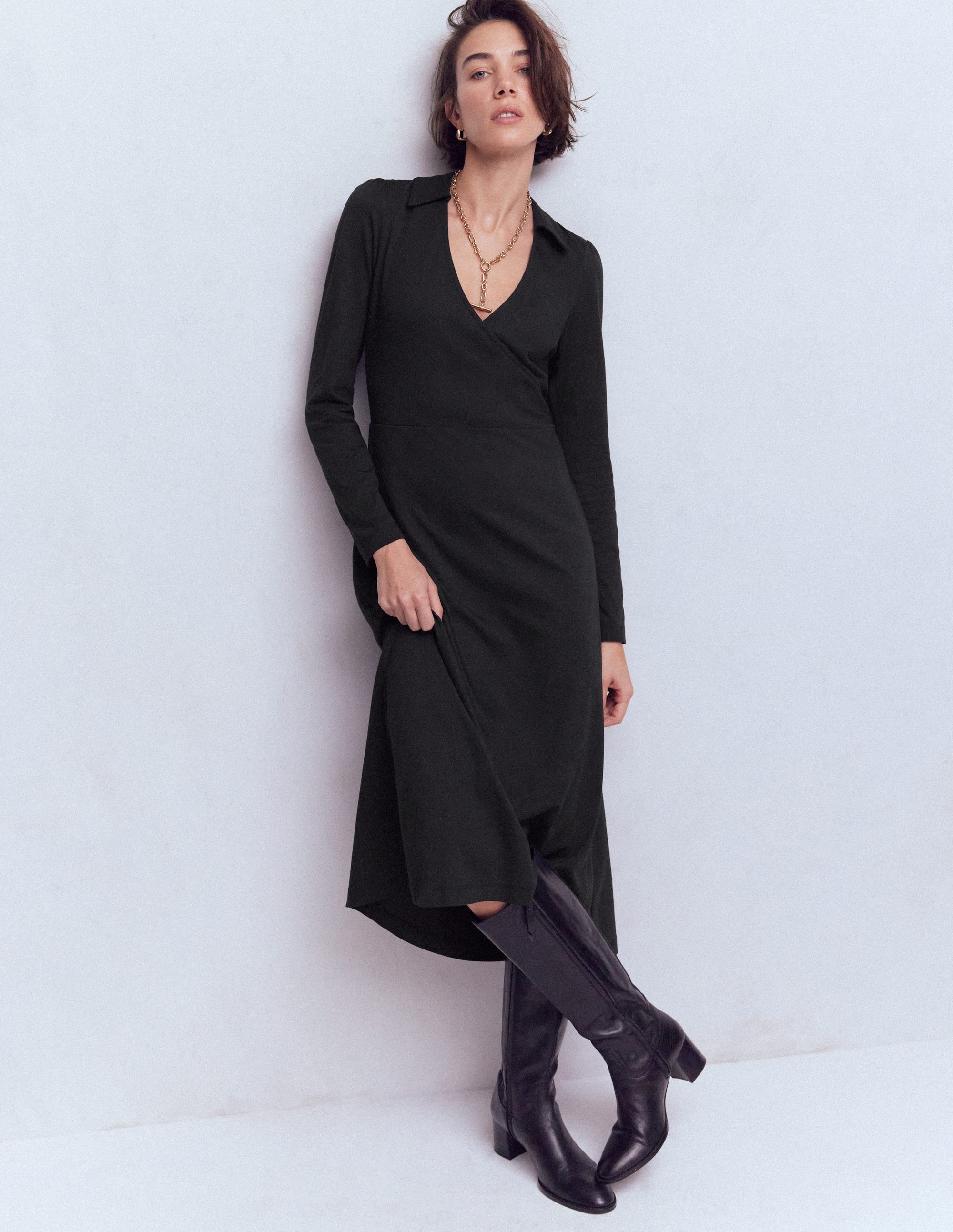A woman wearing a black wrap dress with long sleeves, a V-neckline, and a fitted waist, paired with black knee-high boots.