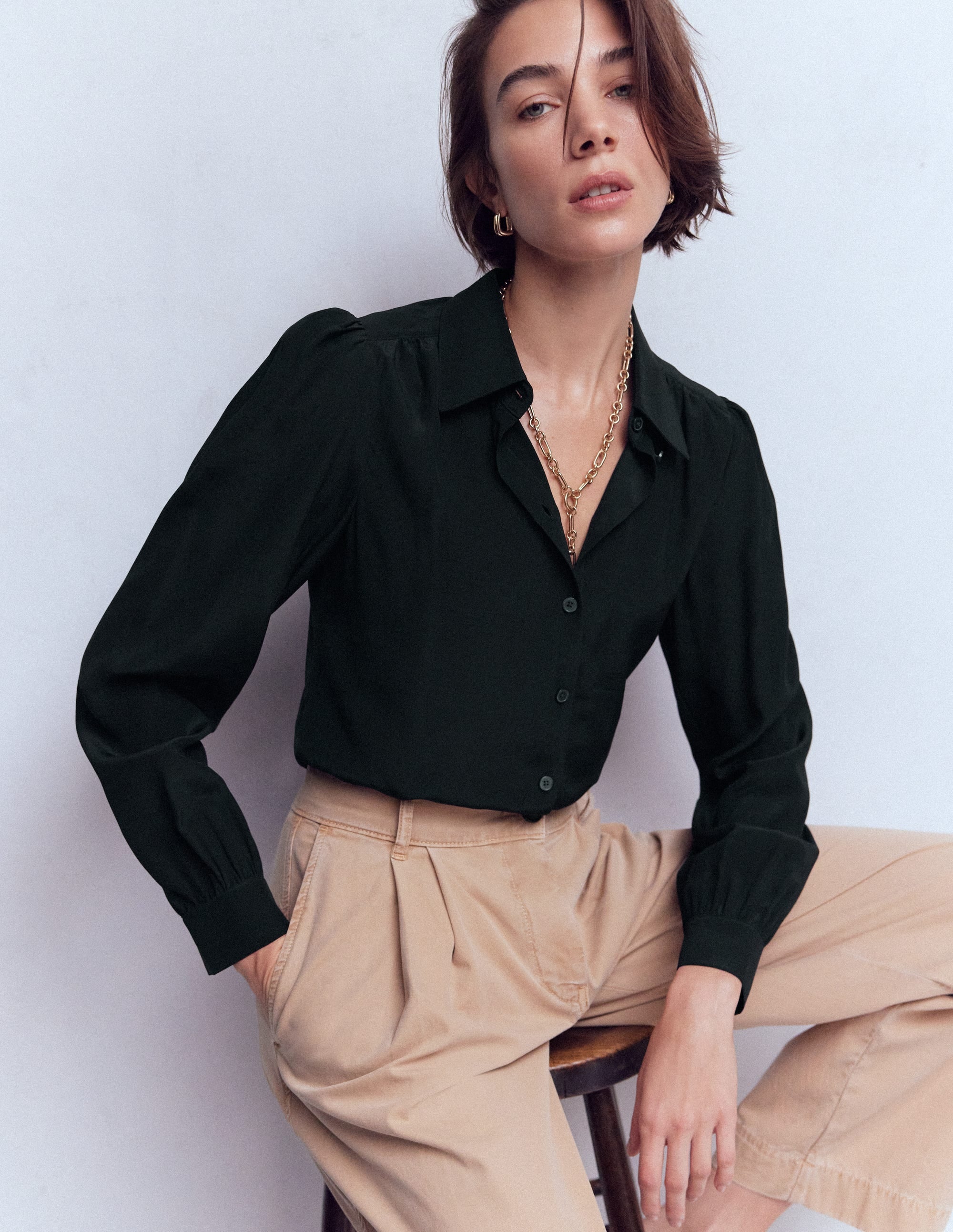 A woman wearing a black silk shirt with a classic collar, button-down front, and long sleeves, paired with beige pants.