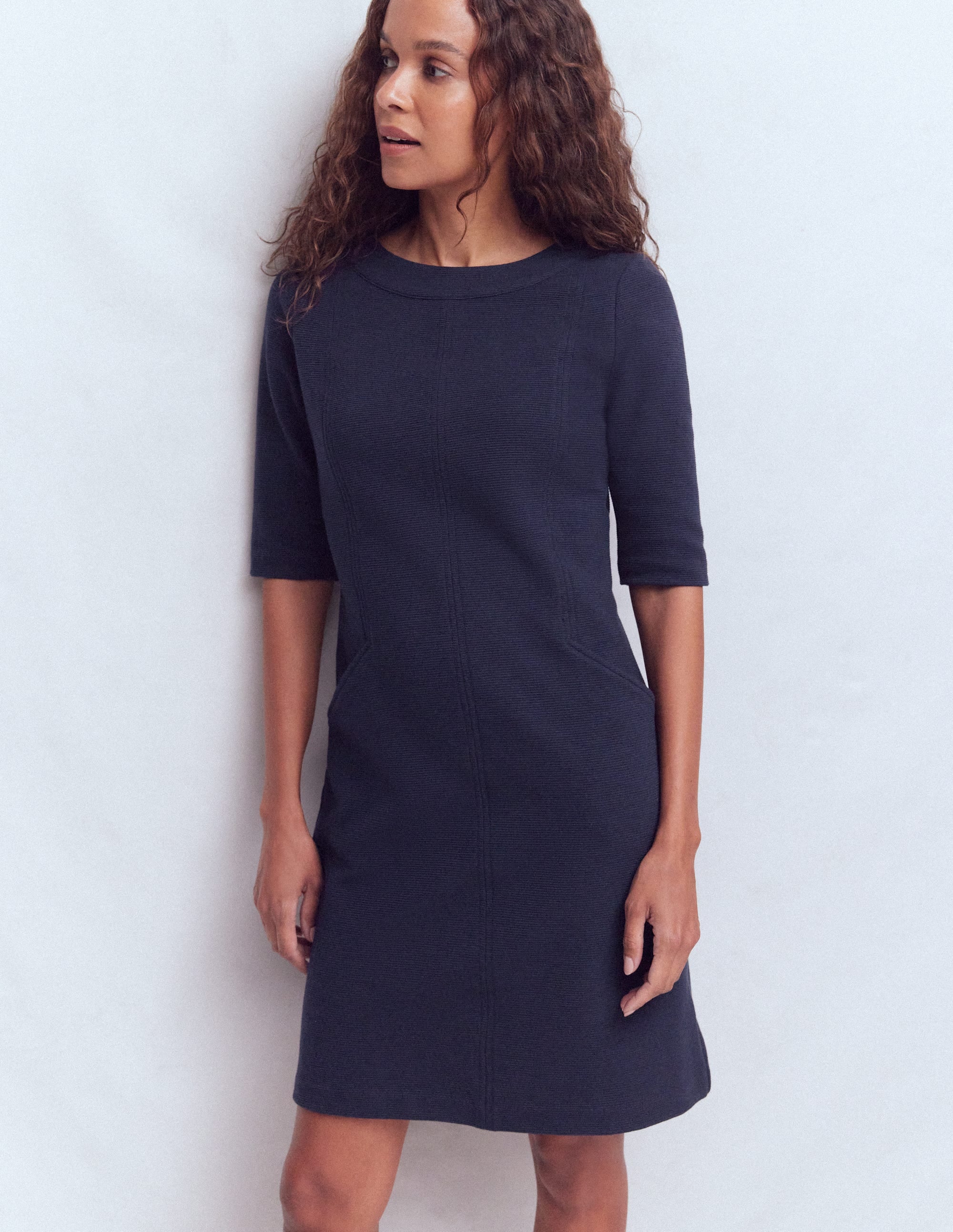 A woman wearing a navy shift dress with a round neckline and three-quarter sleeves, standing against a plain background.