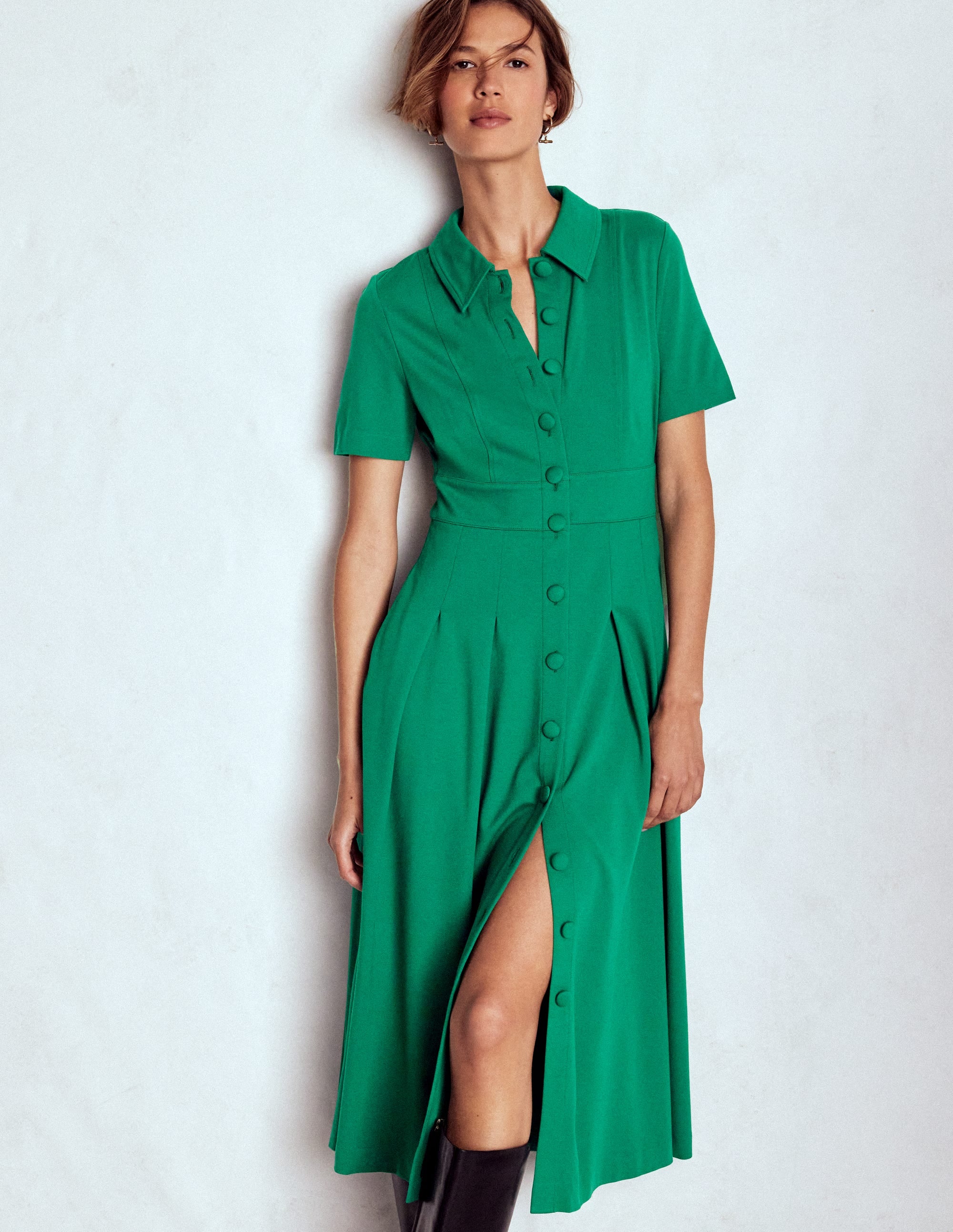 A woman wearing a jewel green shirt dress with short sleeves, fabric-covered buttons, pleats, and pintucks.