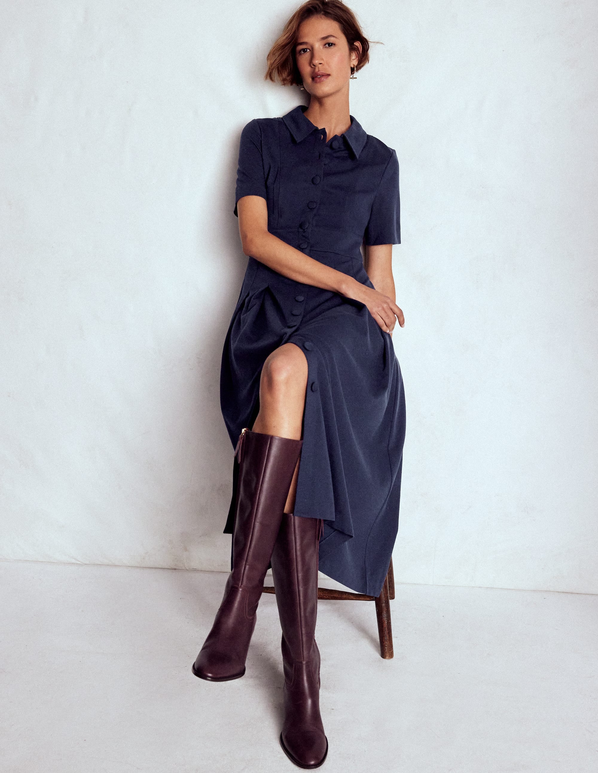 A woman wearing a navy blue shirt dress with short sleeves, fabric-covered buttons, and pleats, paired with tall brown boots.