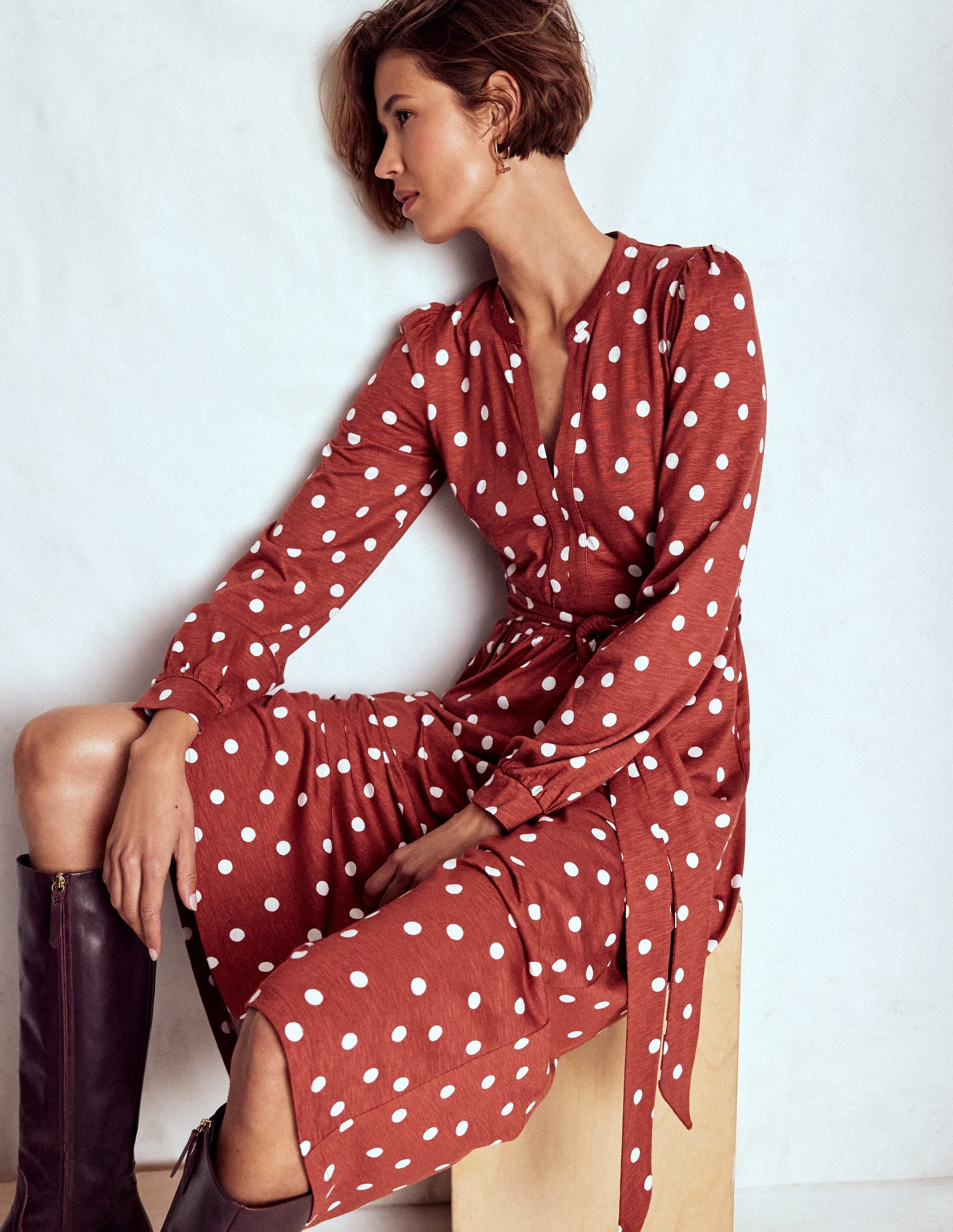 A woman wearing a burnt orange dress with white painted dots, long sleeves, a notch neckline, and a waist tie, sitting on a wooden block.