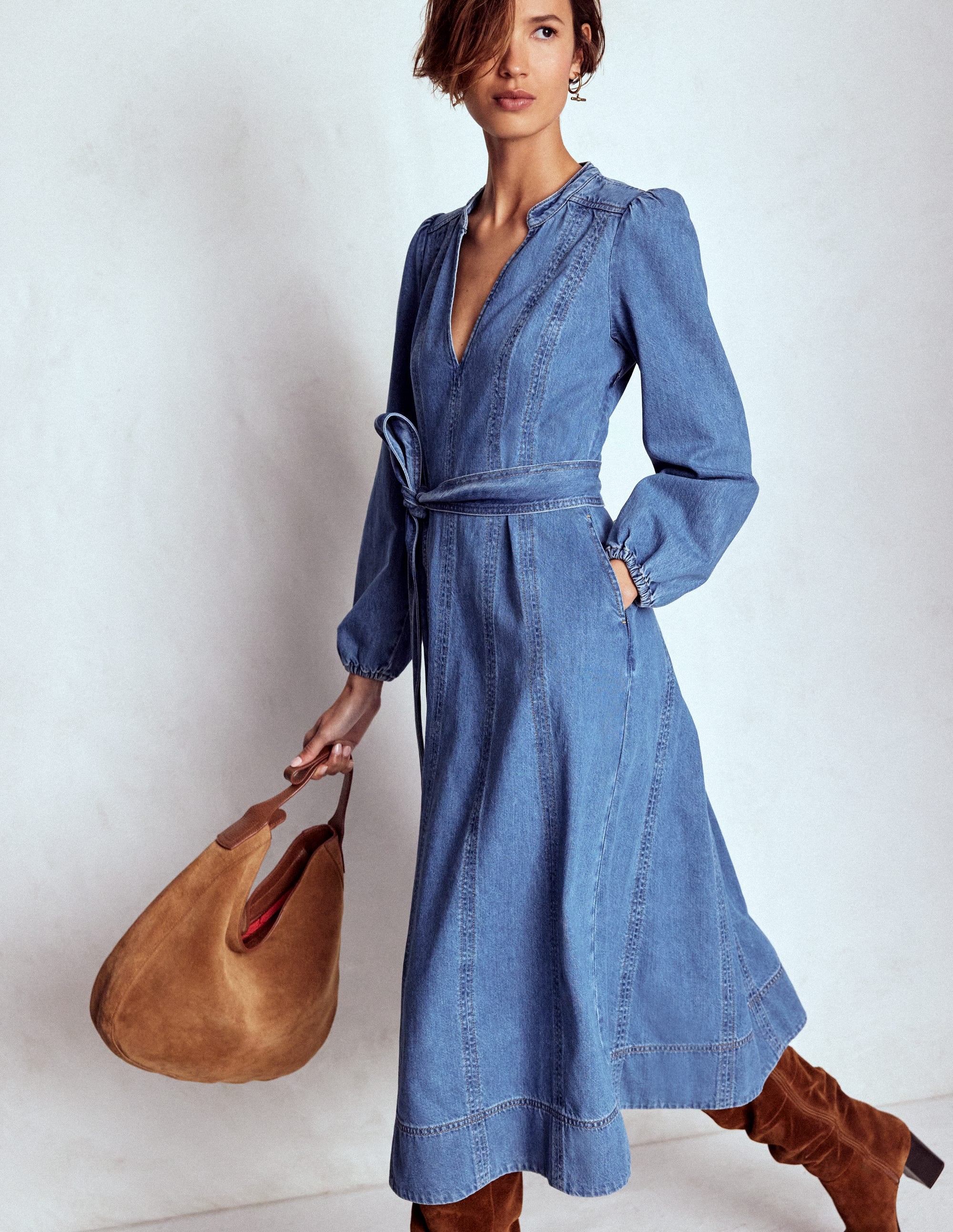 A woman wearing a blue denim flared dress with panel details, notch neckline, long sleeves, and a belt, holding a tan bag.