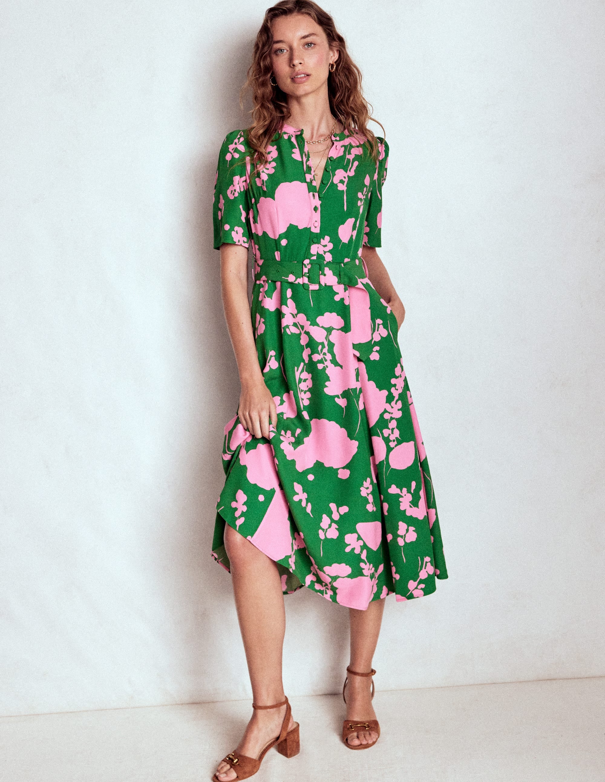 A woman wearing a green dress with pink dainty sprigs pattern, puff sleeves, a belt, and a mid-calf length, standing against a plain background.