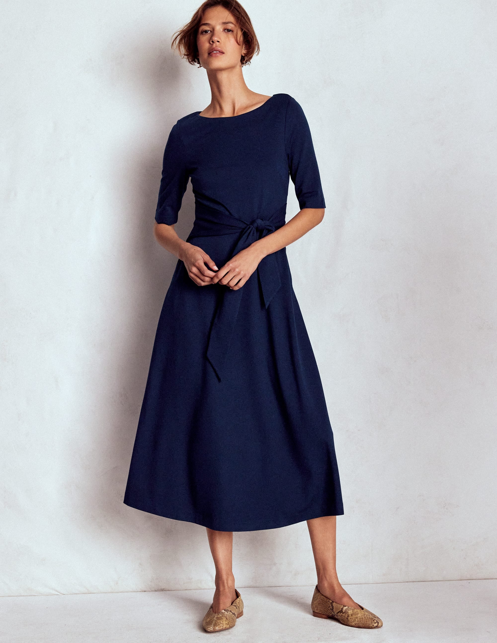 A woman wearing a navy blue fitted dress with elbow-length sleeves, a boat neckline, a front bow at the waistband, and side seam pockets, styled with beige shoes.