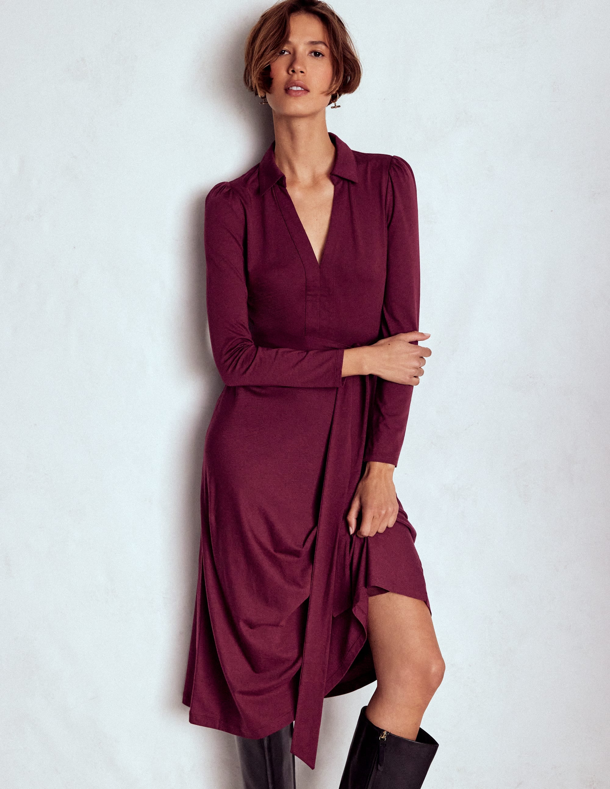 A woman wearing a Deep Wisteria colored, long-sleeved, collared jersey dress with a detachable belt, standing against a plain background.