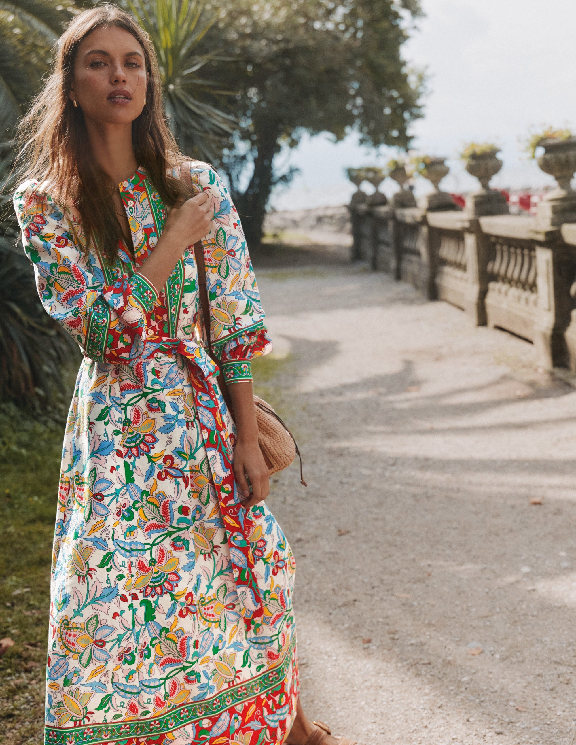 A woman wearing a colorful, patterned midi dress with billowy sleeves and a self-tie belt, standing outdoors near a stone railing with trees and sky in the background.
