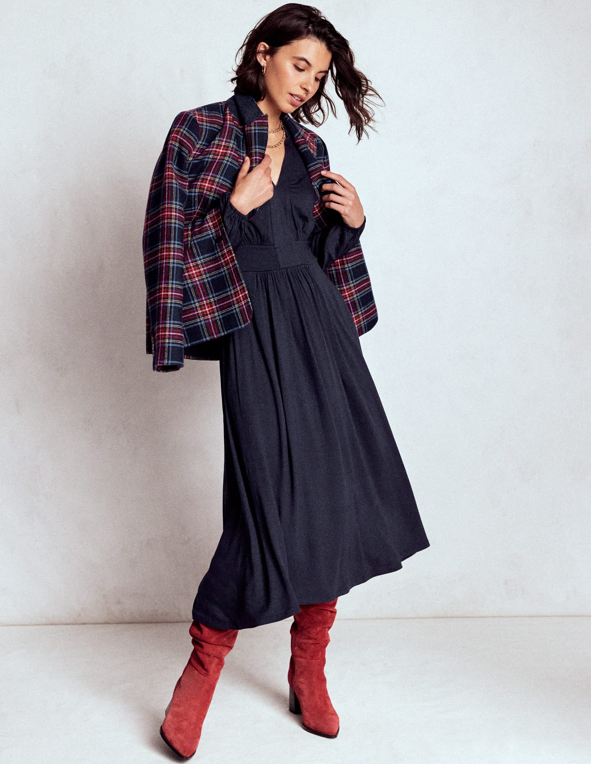 A woman wearing a navy long sleeve midi dress with a V-neckline, contrasted with a plaid jacket, paired with red boots.