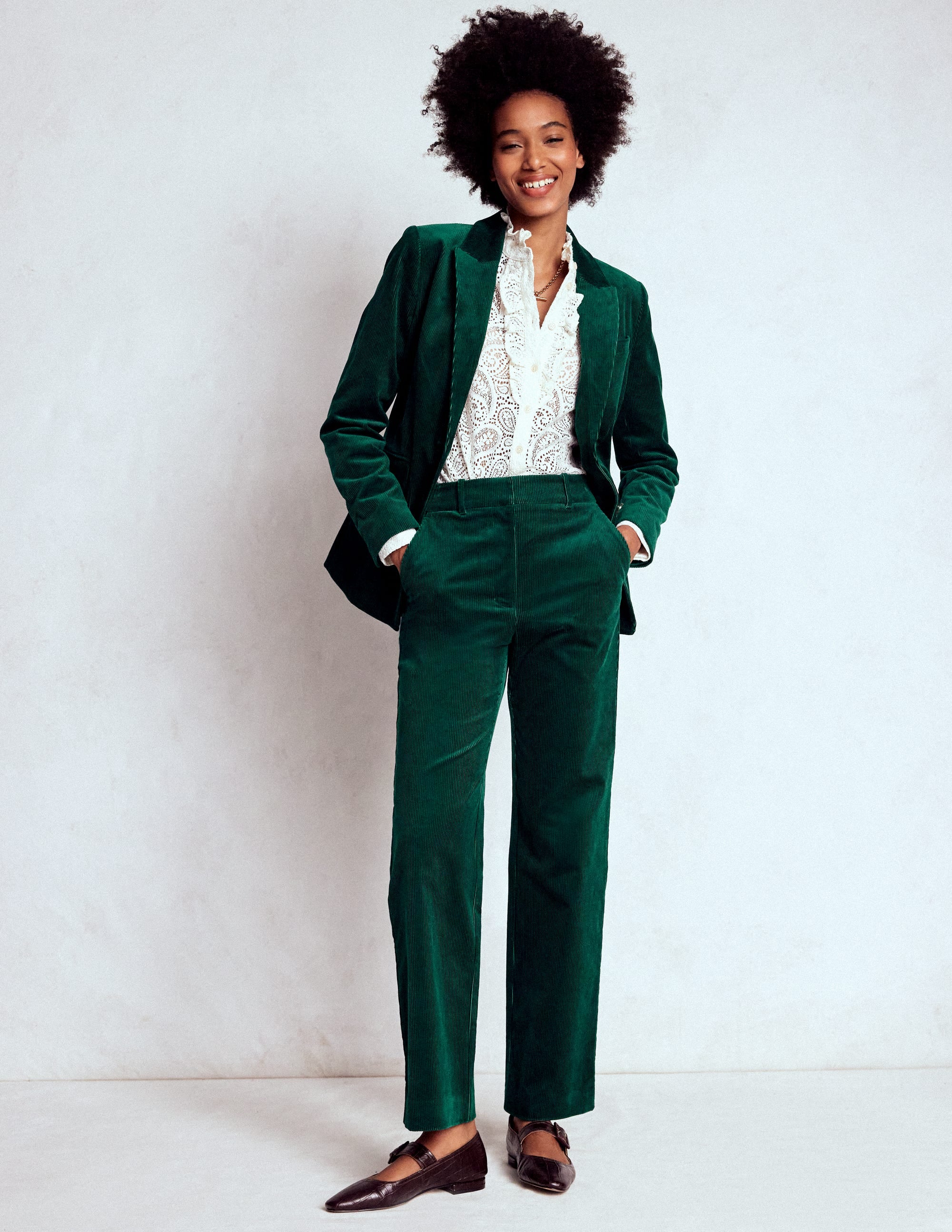 A woman wearing a green corduroy blazer with a classic lapel, pockets, and a structured fit, paired with matching green corduroy pants and a white lace blouse.