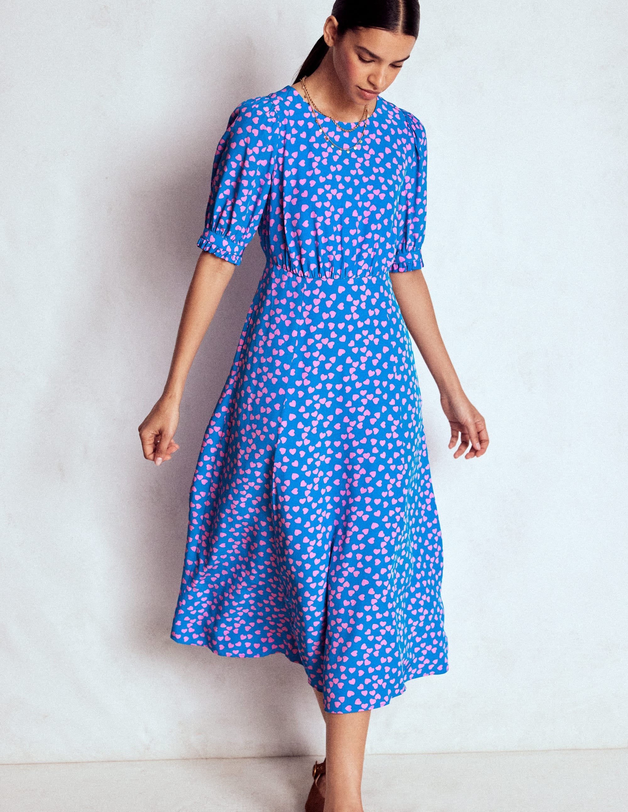 A woman wearing a blue midi tea dress with pink heart pattern, puff sleeves with frilled cuffs, and a full skirt, standing against a plain background.