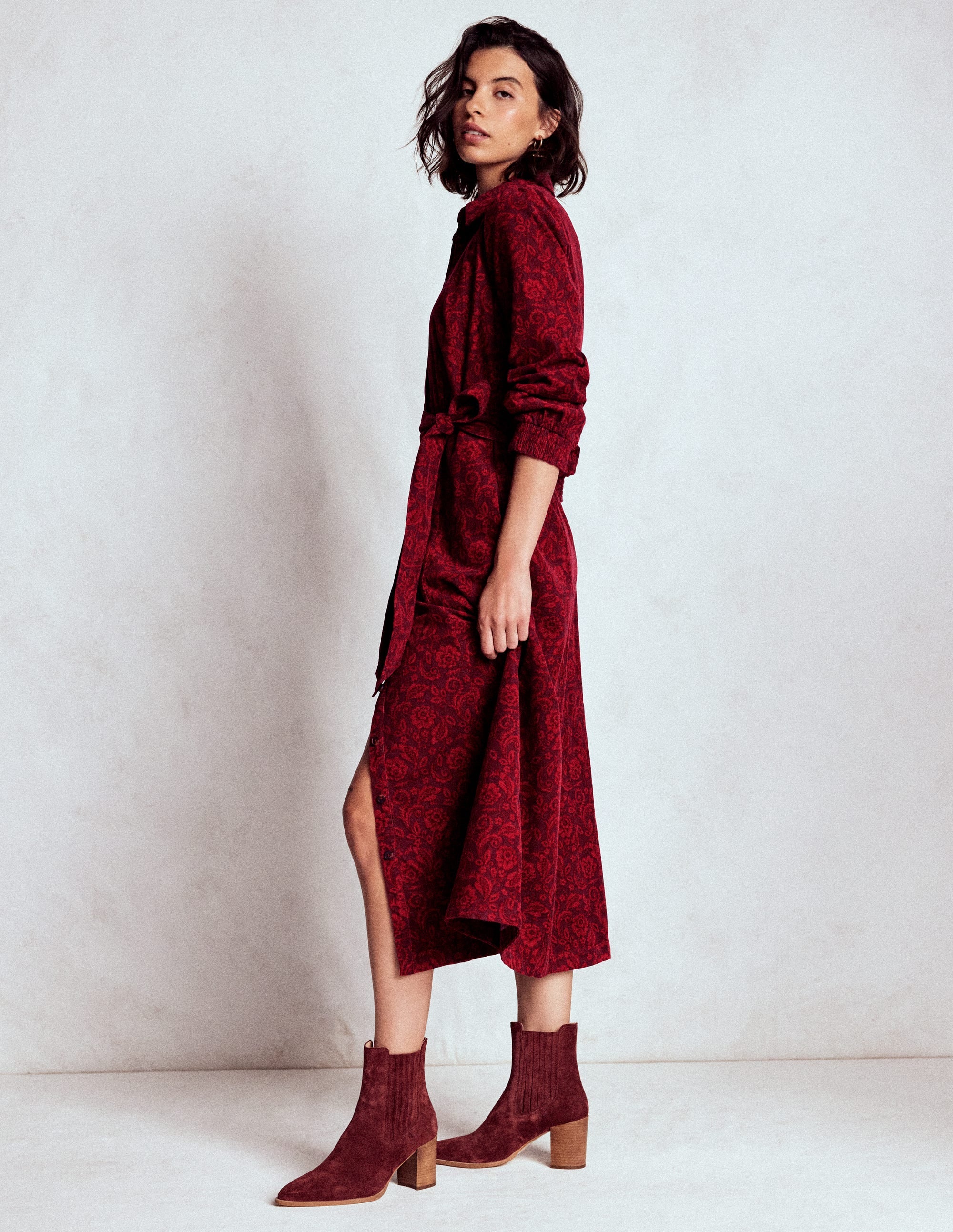 A woman wearing a red floral patterned midi dress with statement sleeves, a self-fabric belt, and ankle boots, standing against a plain background.