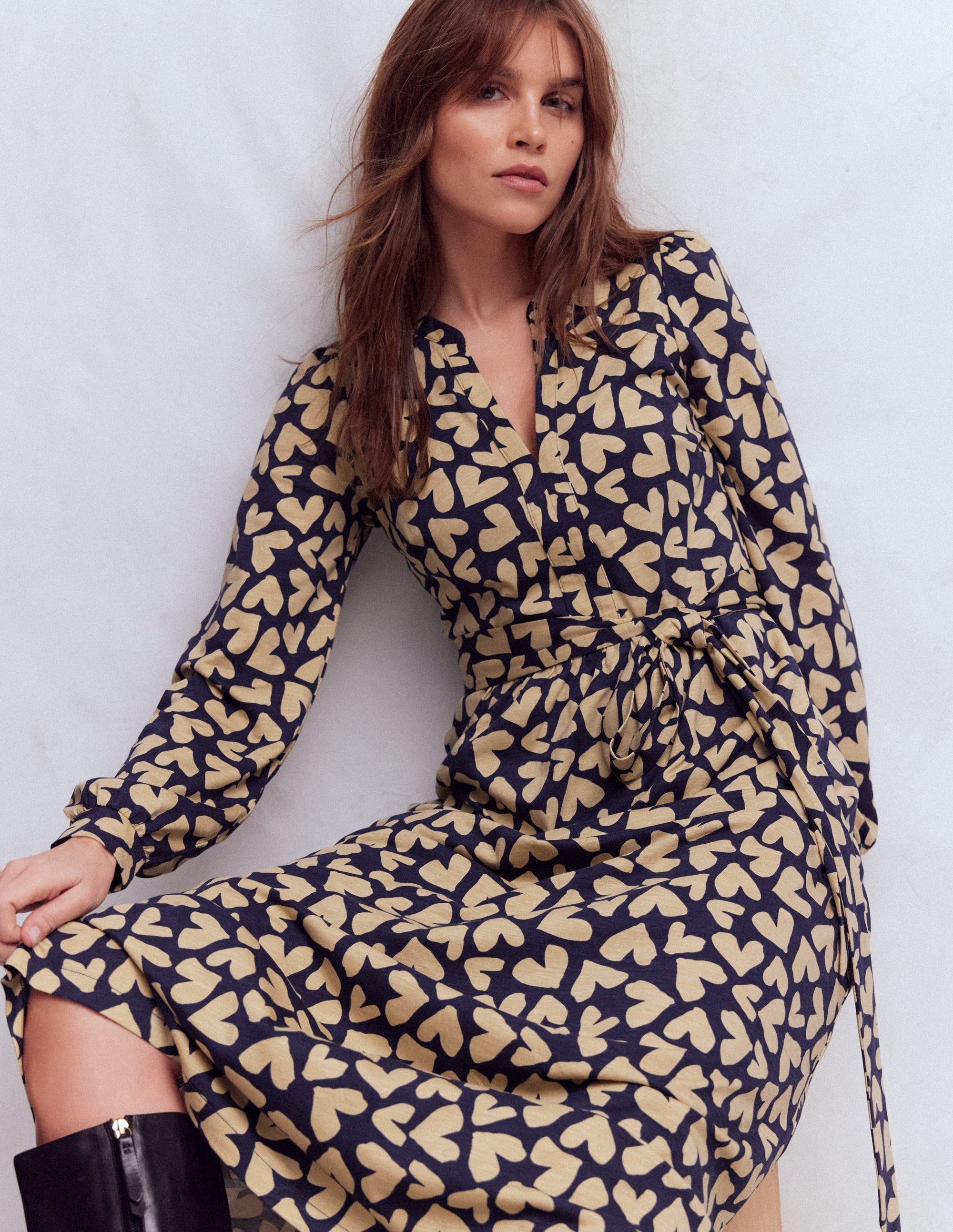 A woman wearing a navy blue dress with a beige heart scatter print, long sleeves, and a self-tie belt.