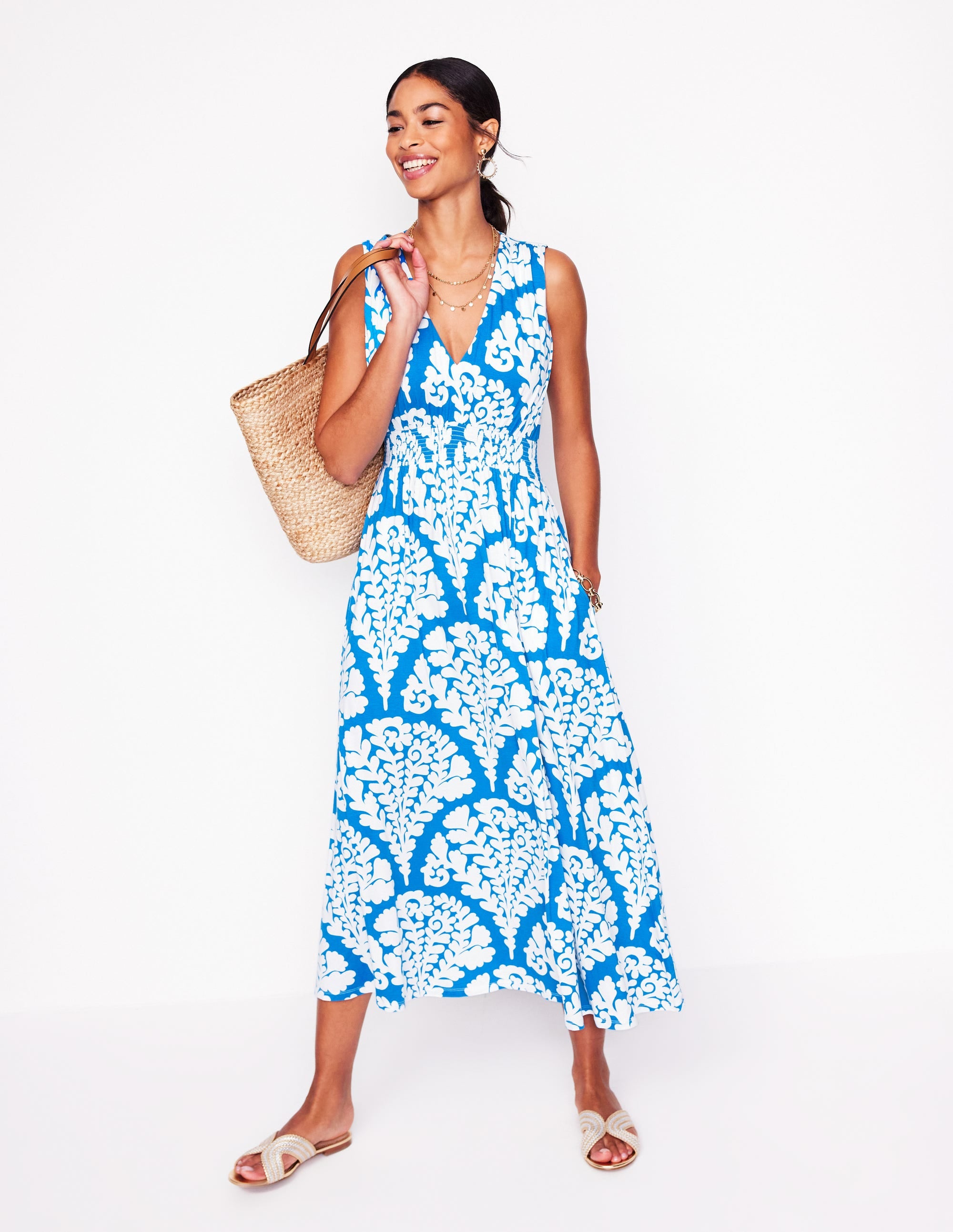 A woman wearing a Cerulean Blue maxi dress with bright floral prints, sleeveless design, and a V-neckline, accessorized with jewelry and a straw bag.