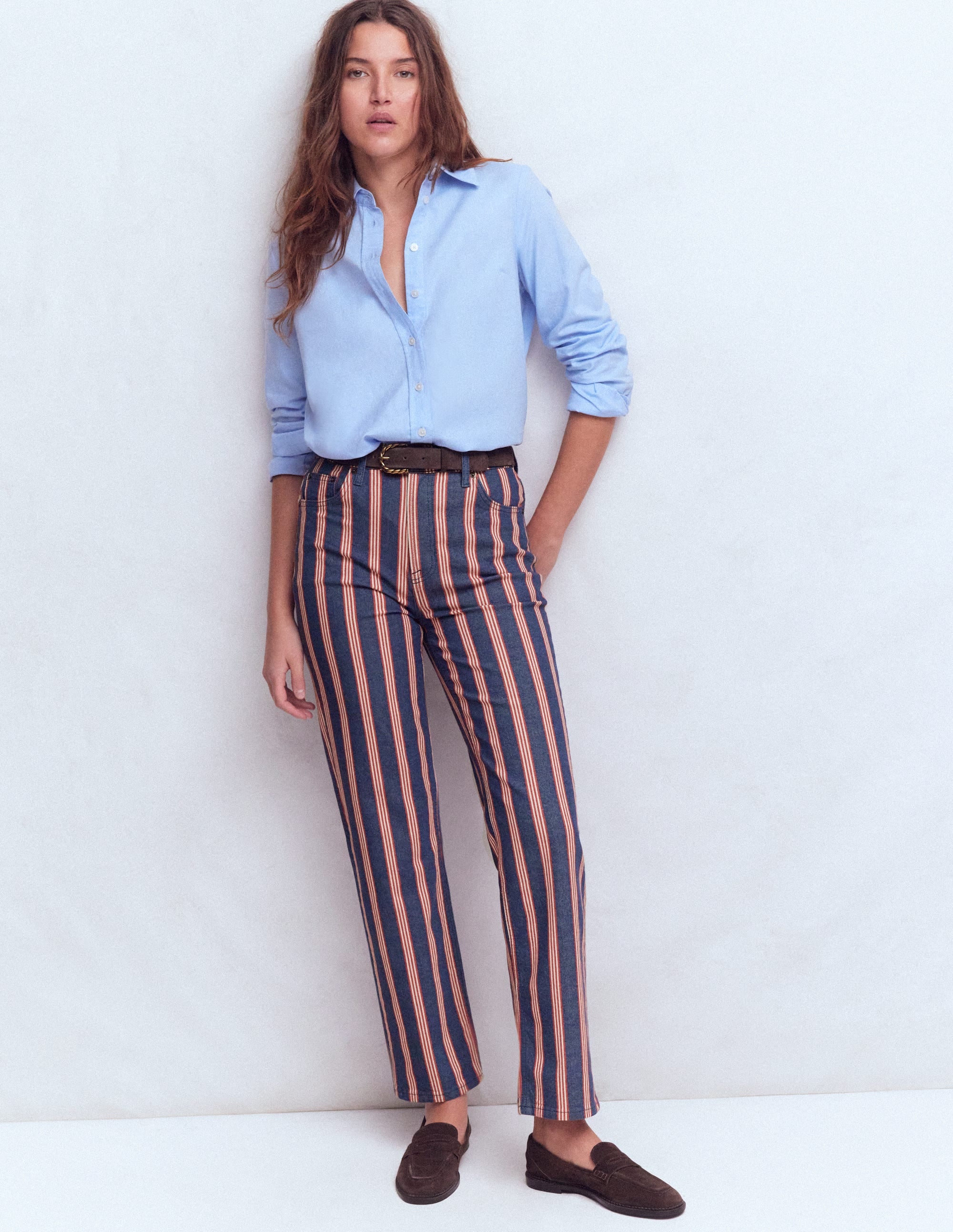 A woman wearing navy high rise straight jeans with red and white vertical stripes, paired with a light blue button-up shirt and dark loafers.
