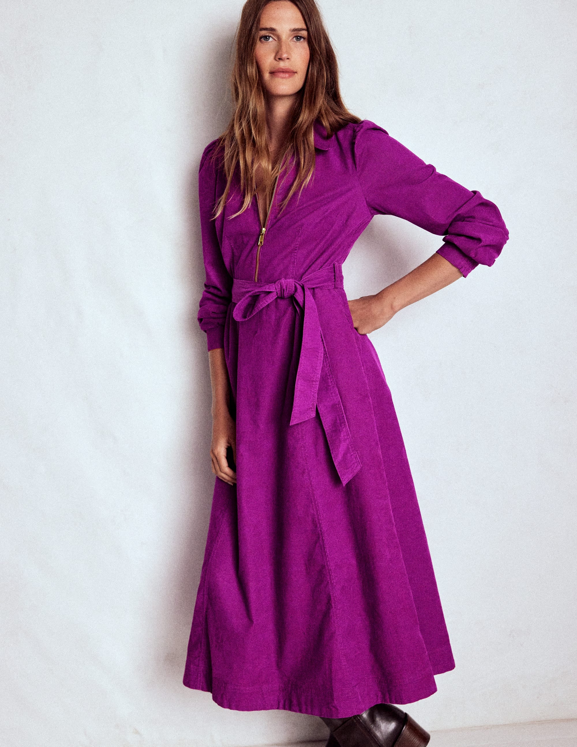 A woman wearing a regal purple midi dress with a zip-up top, fabric belt, and pockets, standing against a plain background.
