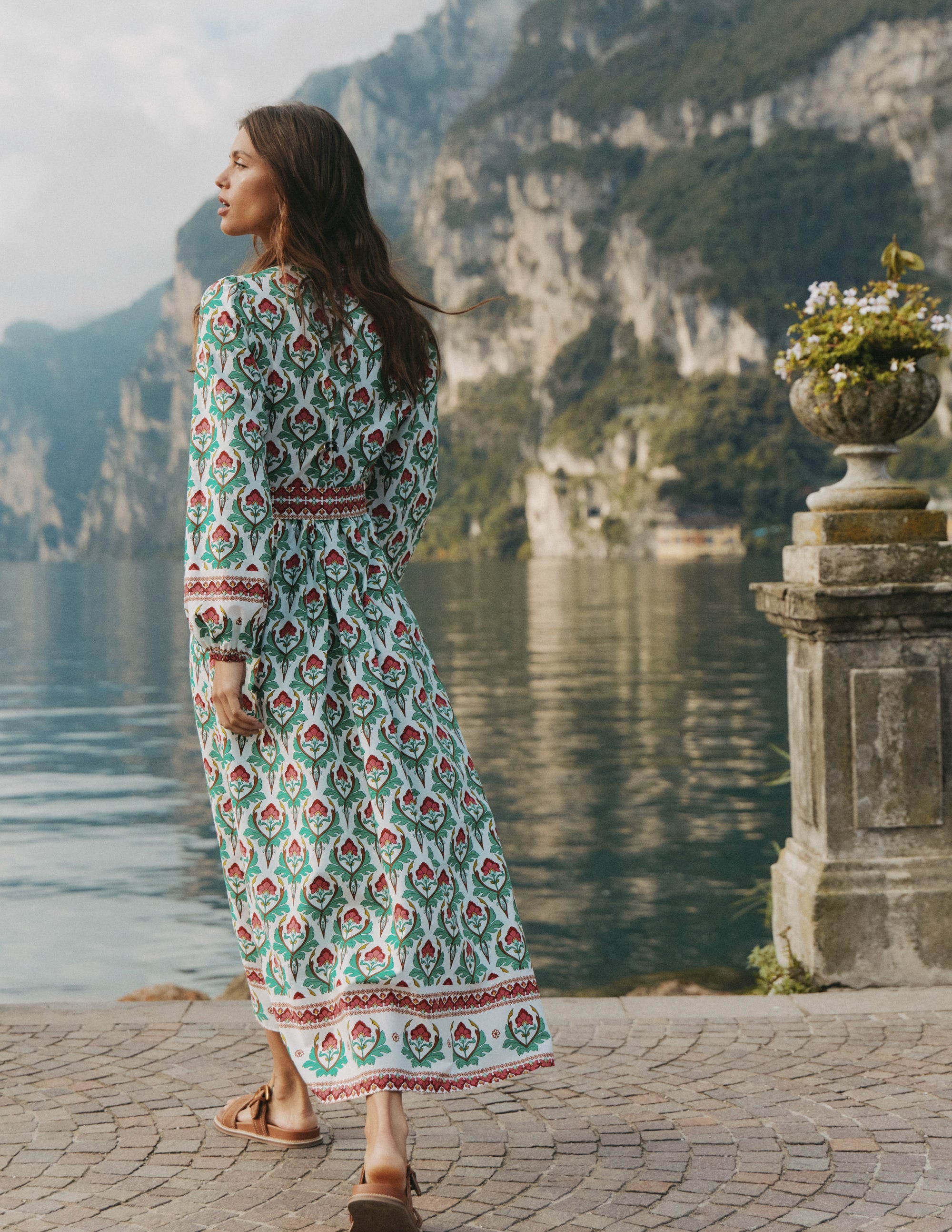 A woman wearing a multicolour patterned midi dress with long sleeves and a self-tie belt, standing near a body of water with mountains in the background.