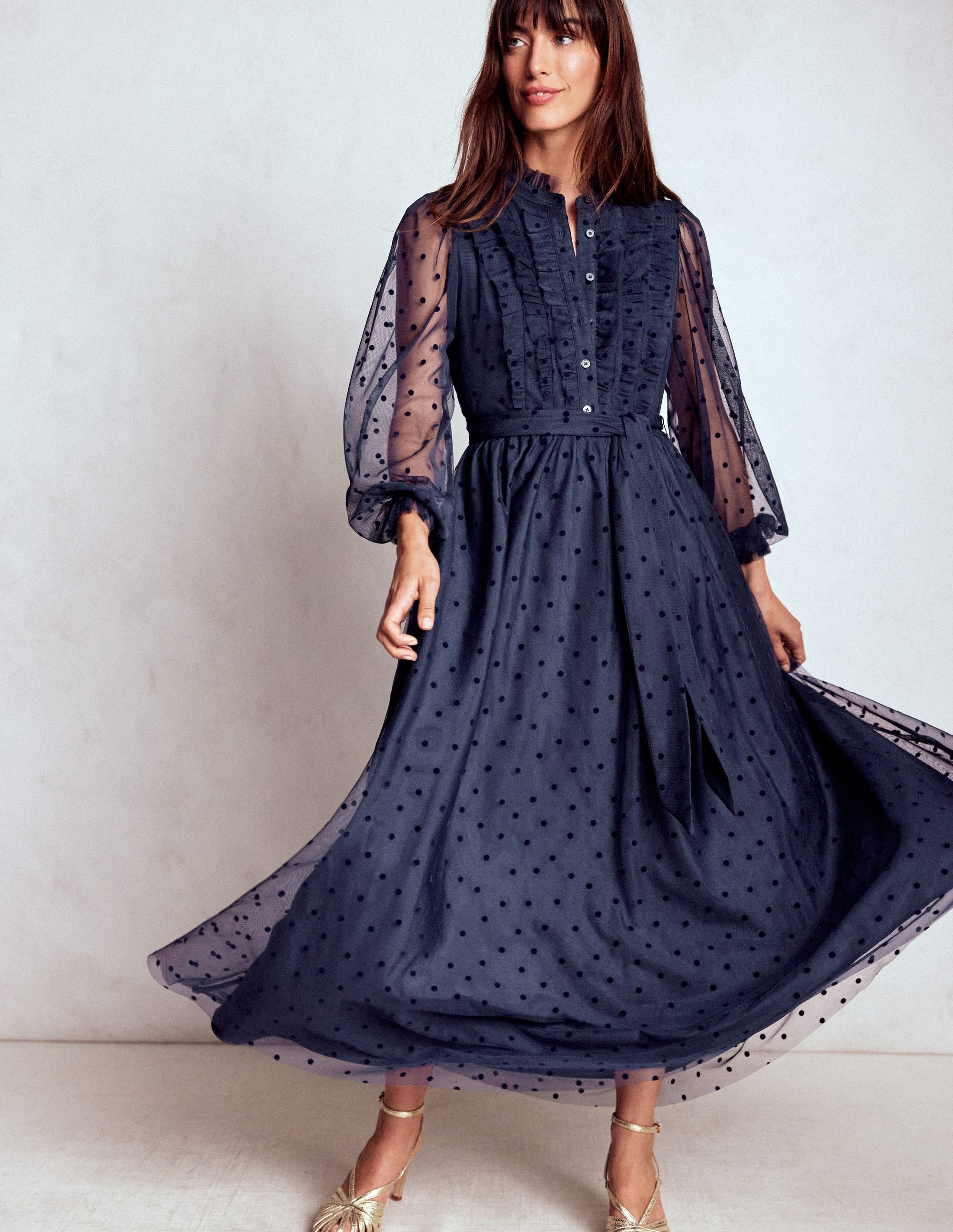 A woman wearing a navy blue maxi dress with ruffles, sheer polka dot sleeves, and a flowing skirt.