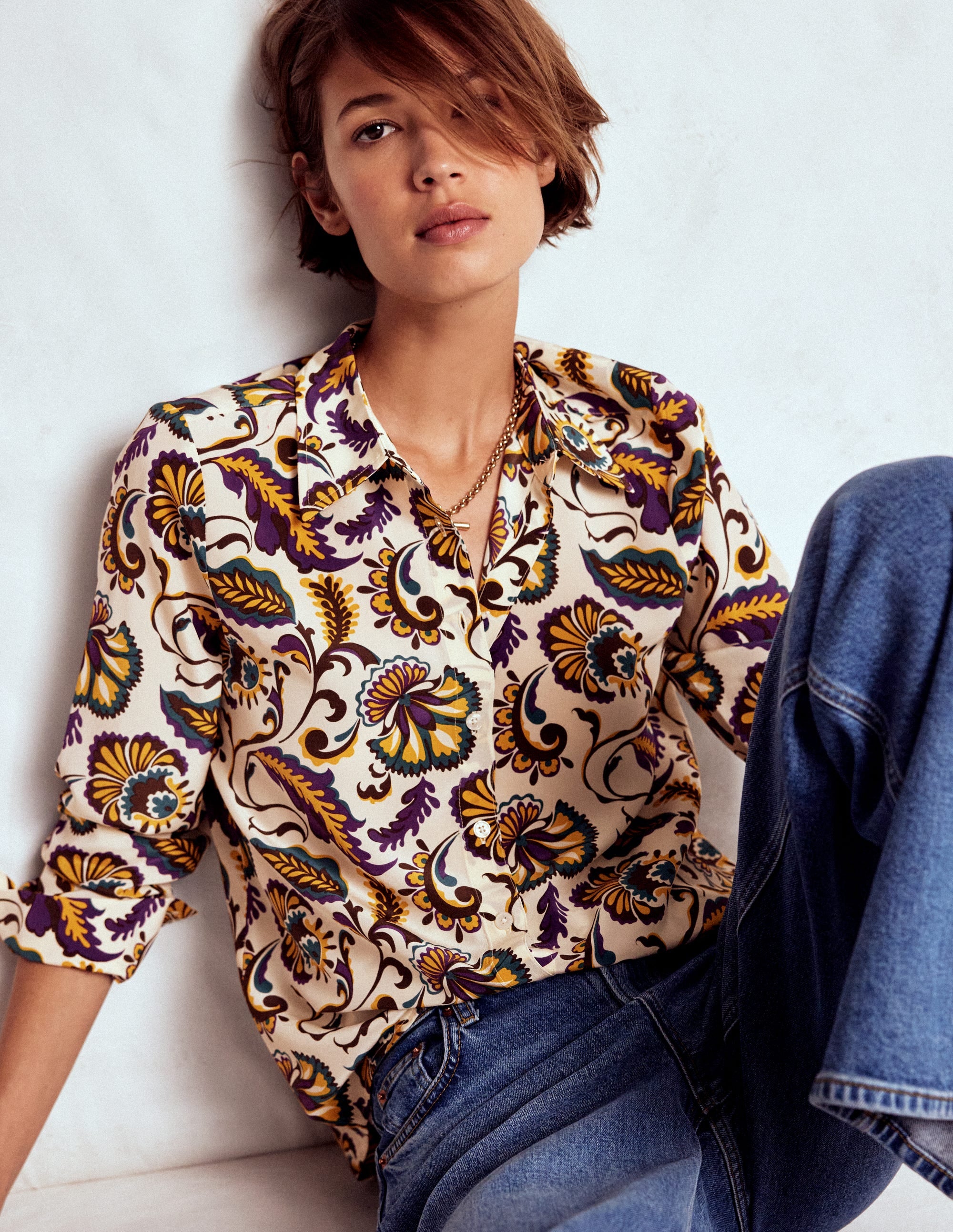 A woman wearing a silk shirt with a multicolour ornate meadow print, featuring purple, yellow, blue, and black colors, with a collar and button-down front.
