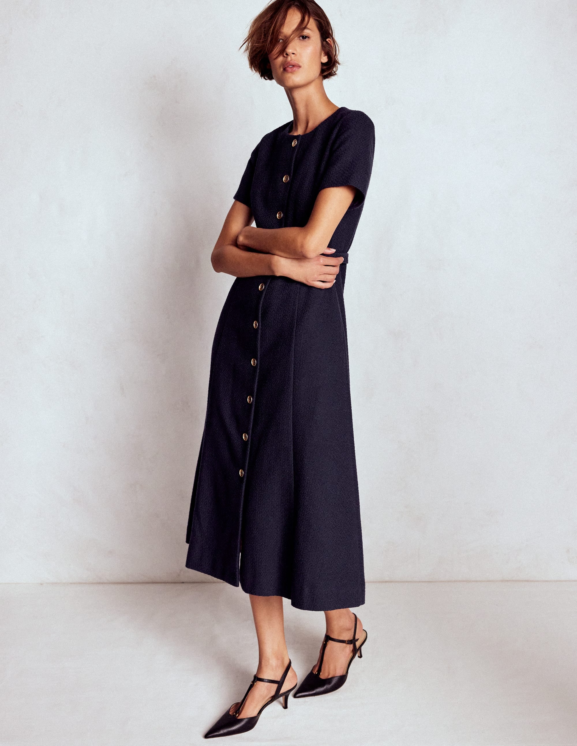 A woman wearing a navy midi dress with short sleeves, buttons down the front, and a fabric belt, paired with black high heels.