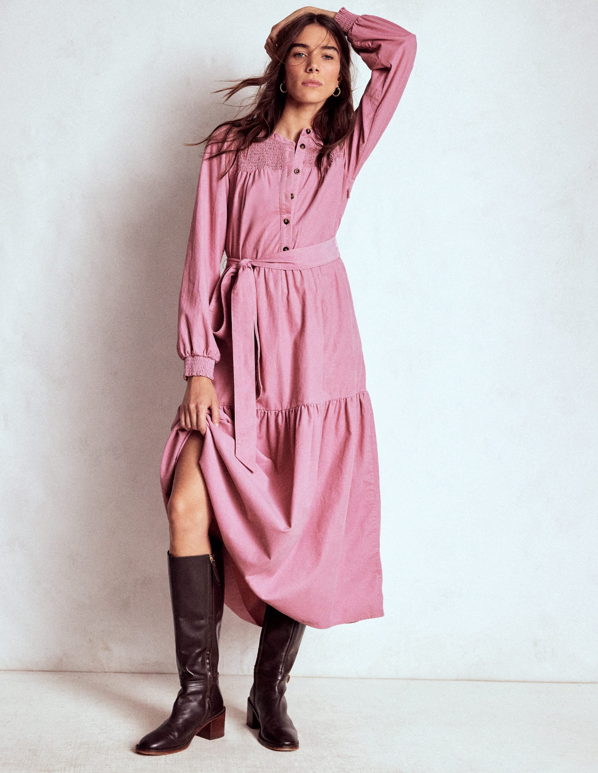A woman wearing a pink midi dress with long sleeves, smocking detail, and a gathered tiered skirt, paired with black knee-high boots.