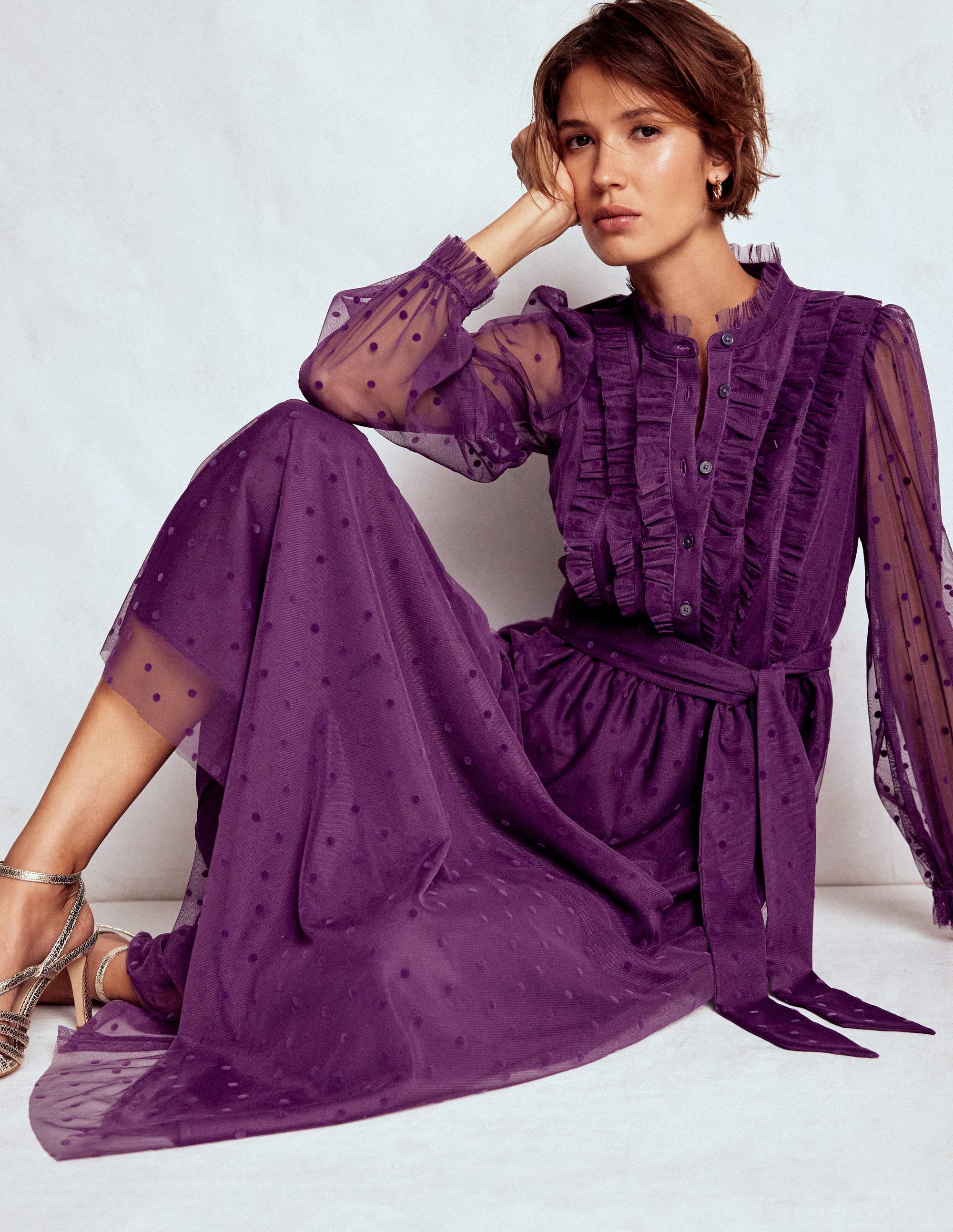 A woman wearing a Viola Purple tulle maxi dress with ruffles, sheer sleeves, and a spot print, sitting against a plain background.