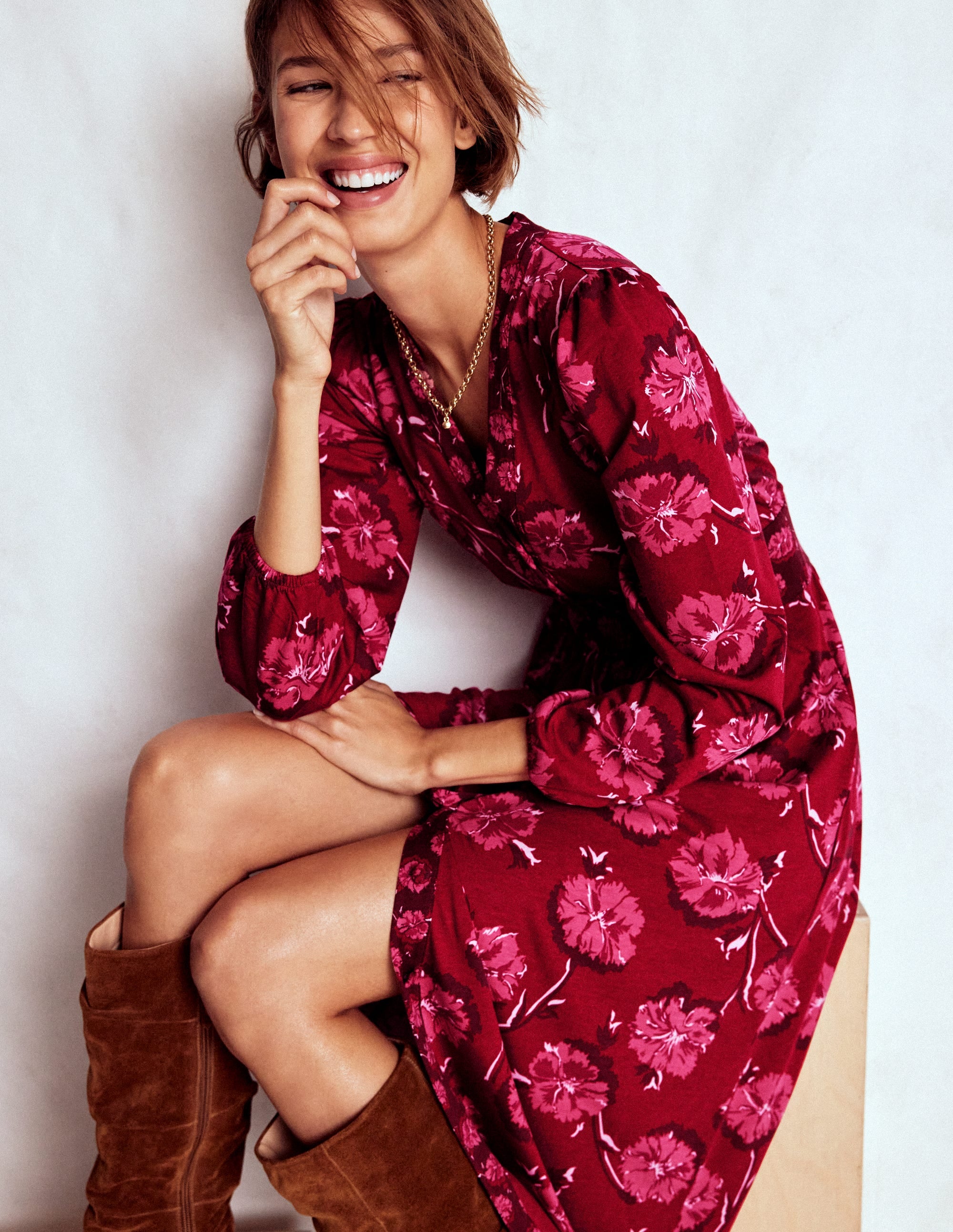 A woman wearing a red long sleeve tea dress with a floral pattern, smiling and sitting with one leg crossed, paired with brown boots.