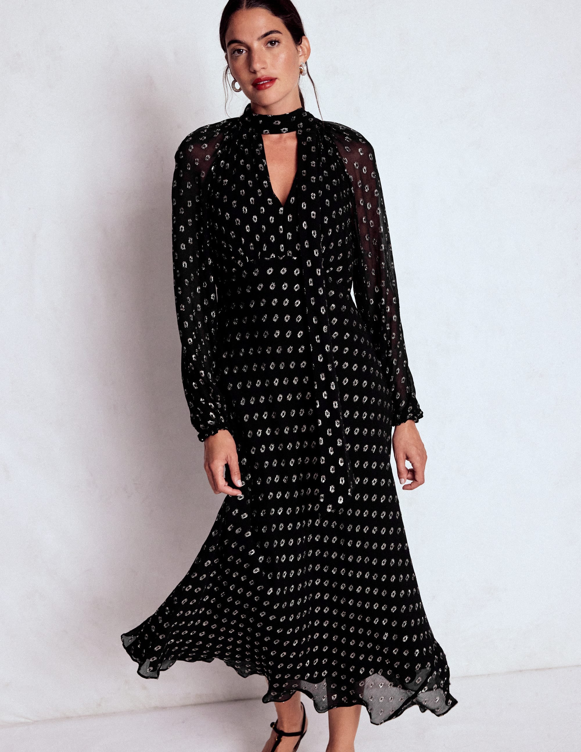 A woman wearing a black midi dress with a tie neck, sheer long sleeves, and a fit and flare silhouette.