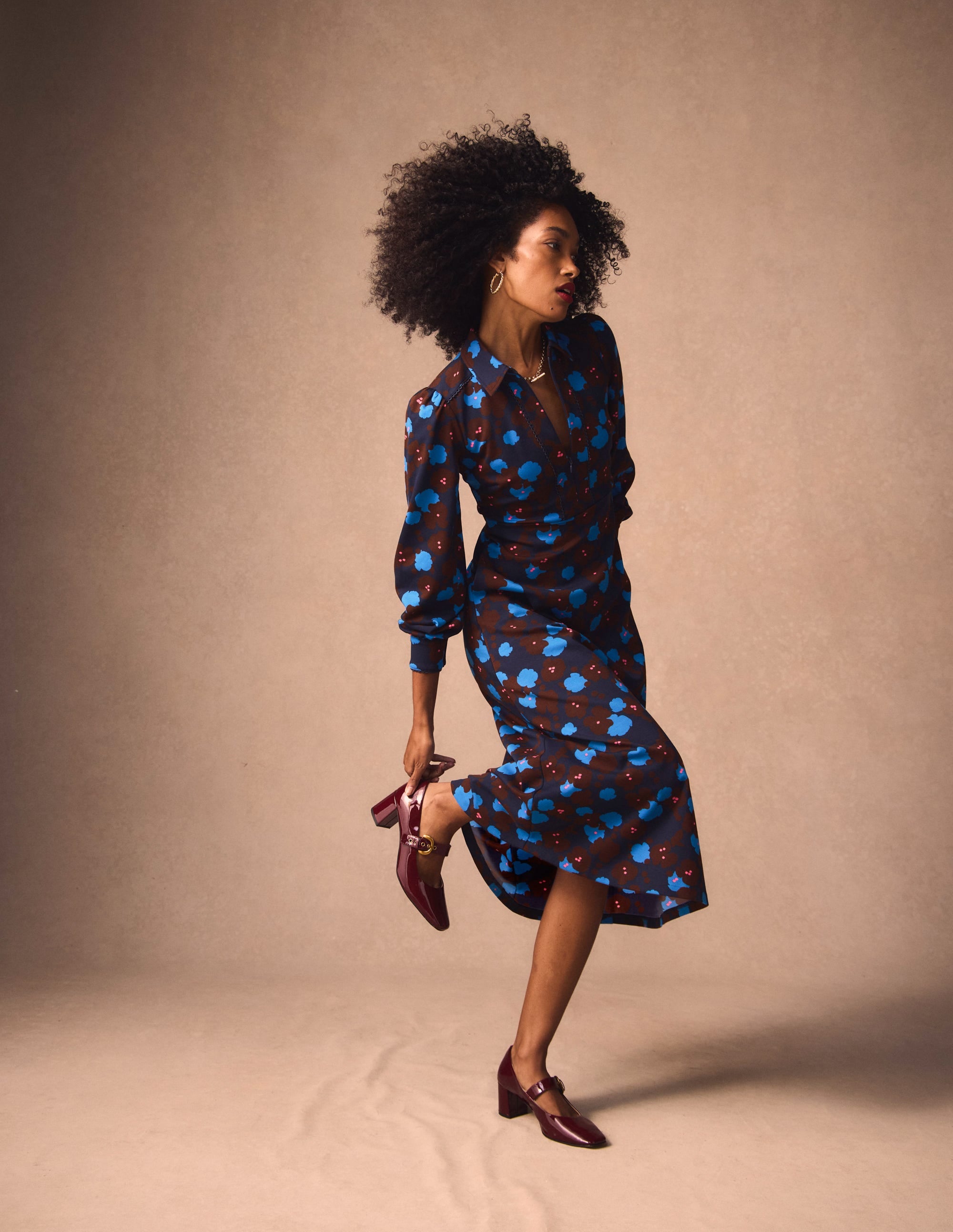 A woman wearing a navy blue midi dress with a floral pattern, long sleeves with buttoned cuffs, and a collar, standing against a plain background.
