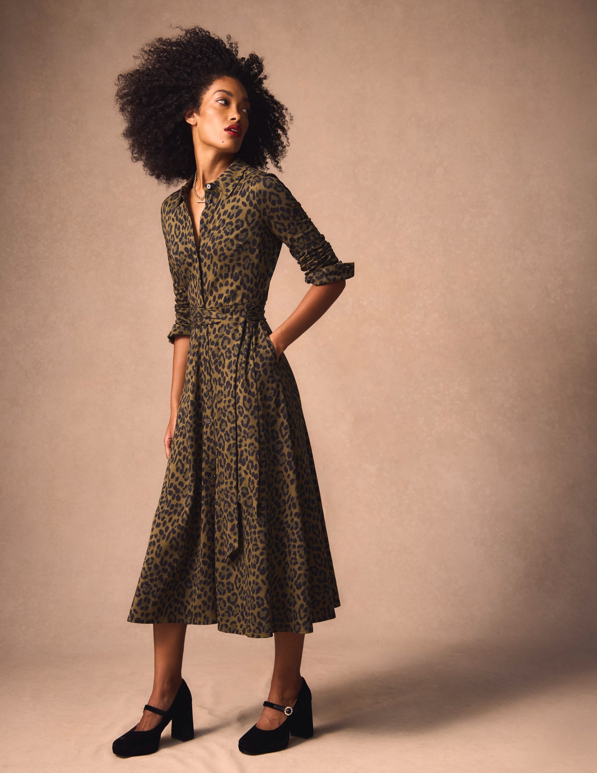 A woman wearing a military olive-colored shirt dress with a brushed animal print, long sleeves, and a waist belt, paired with black high heels.