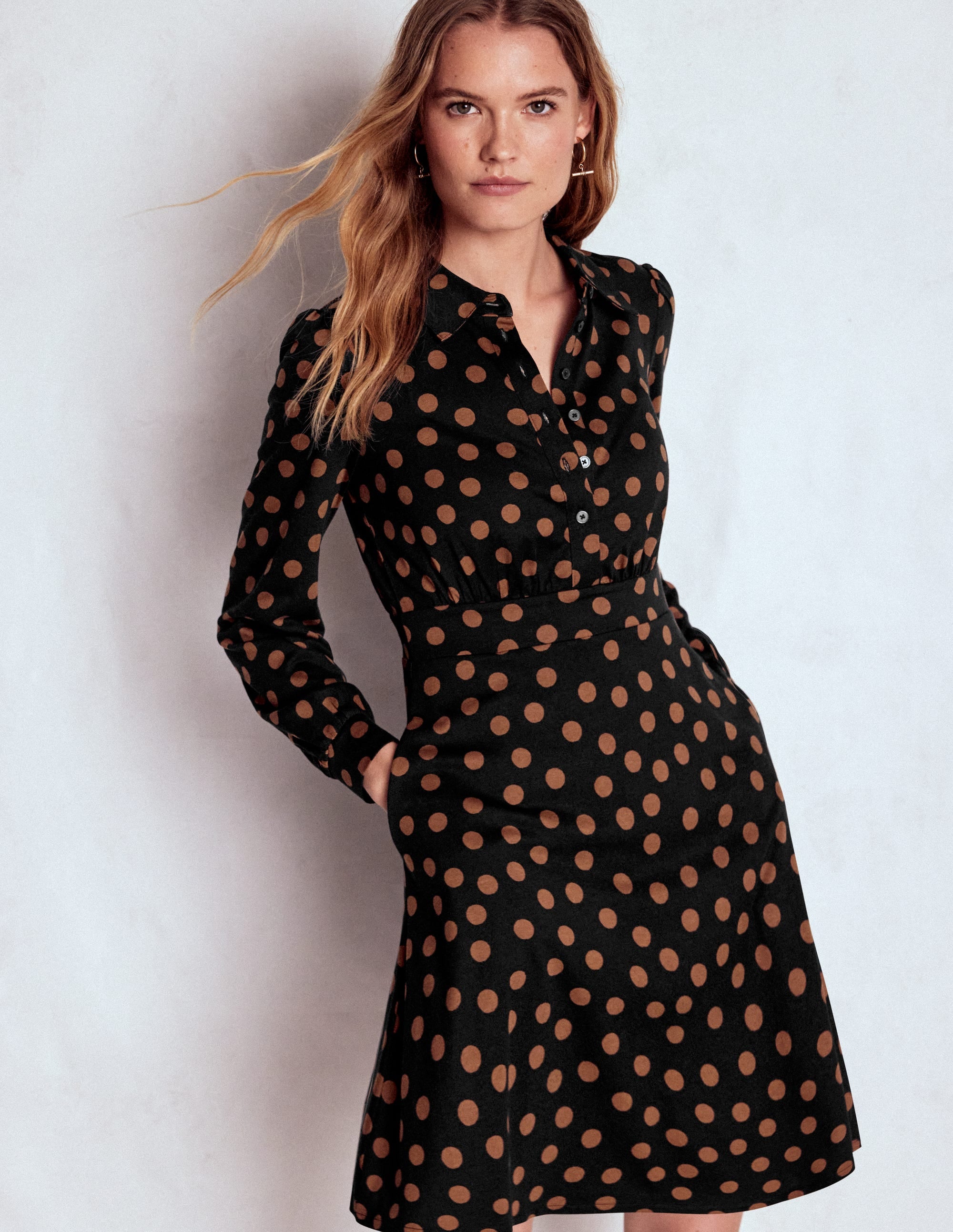 A woman wearing a black shirt dress with rust-colored painted dots, long sleeves, and a button-up front, standing against a plain background.