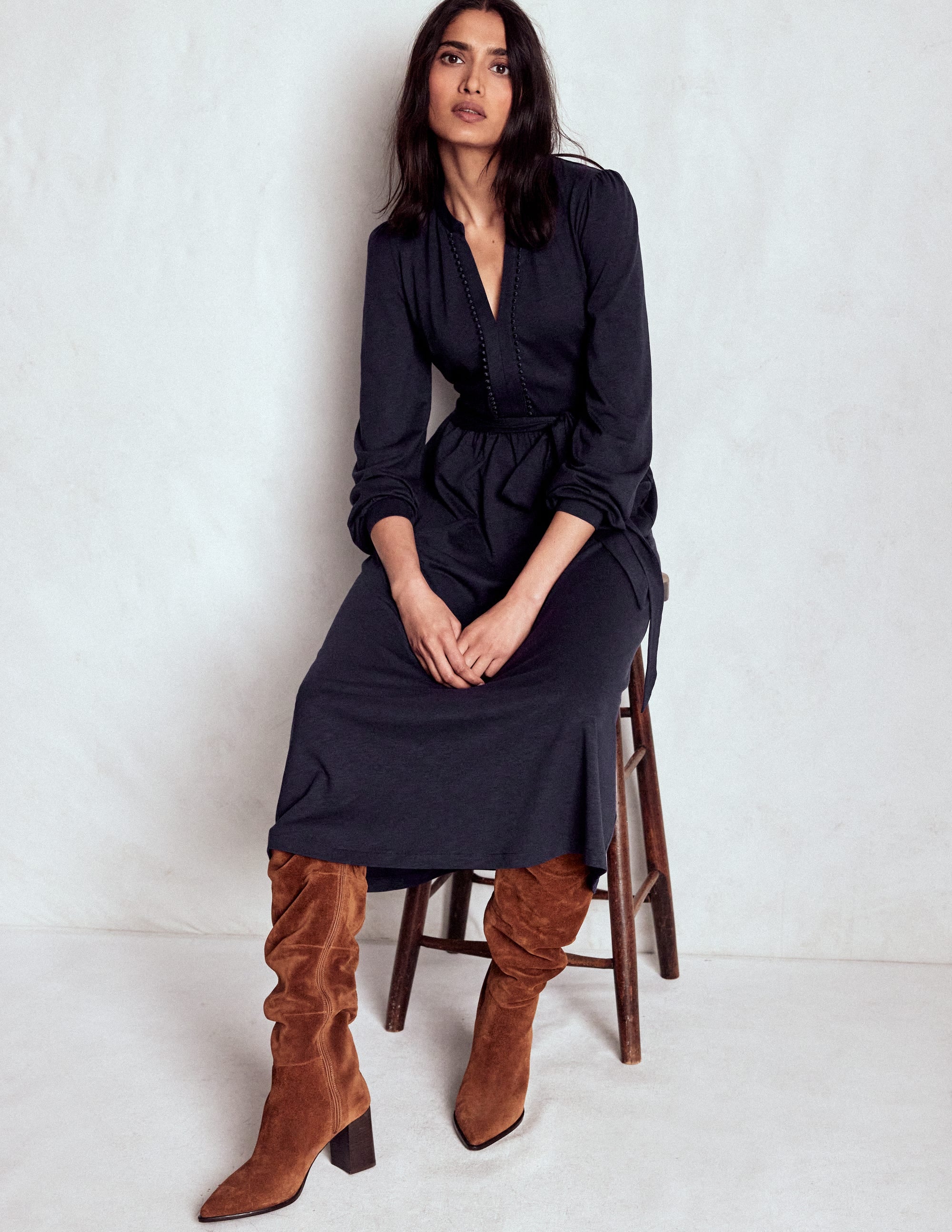 A woman wearing a navy long sleeve jersey dress with a self-tie belt, deep pockets, and a fit and flare silhouette, sitting on a wooden chair.