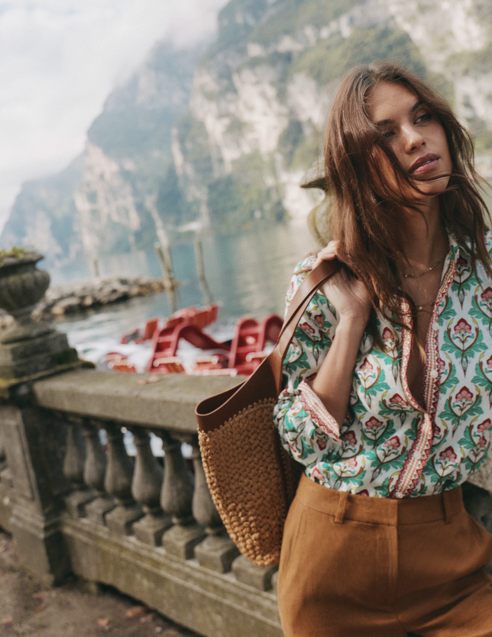 A woman wearing a multi-colored patterned linen shirt, standing outdoors near a stone railing with a scenic water and mountain background.