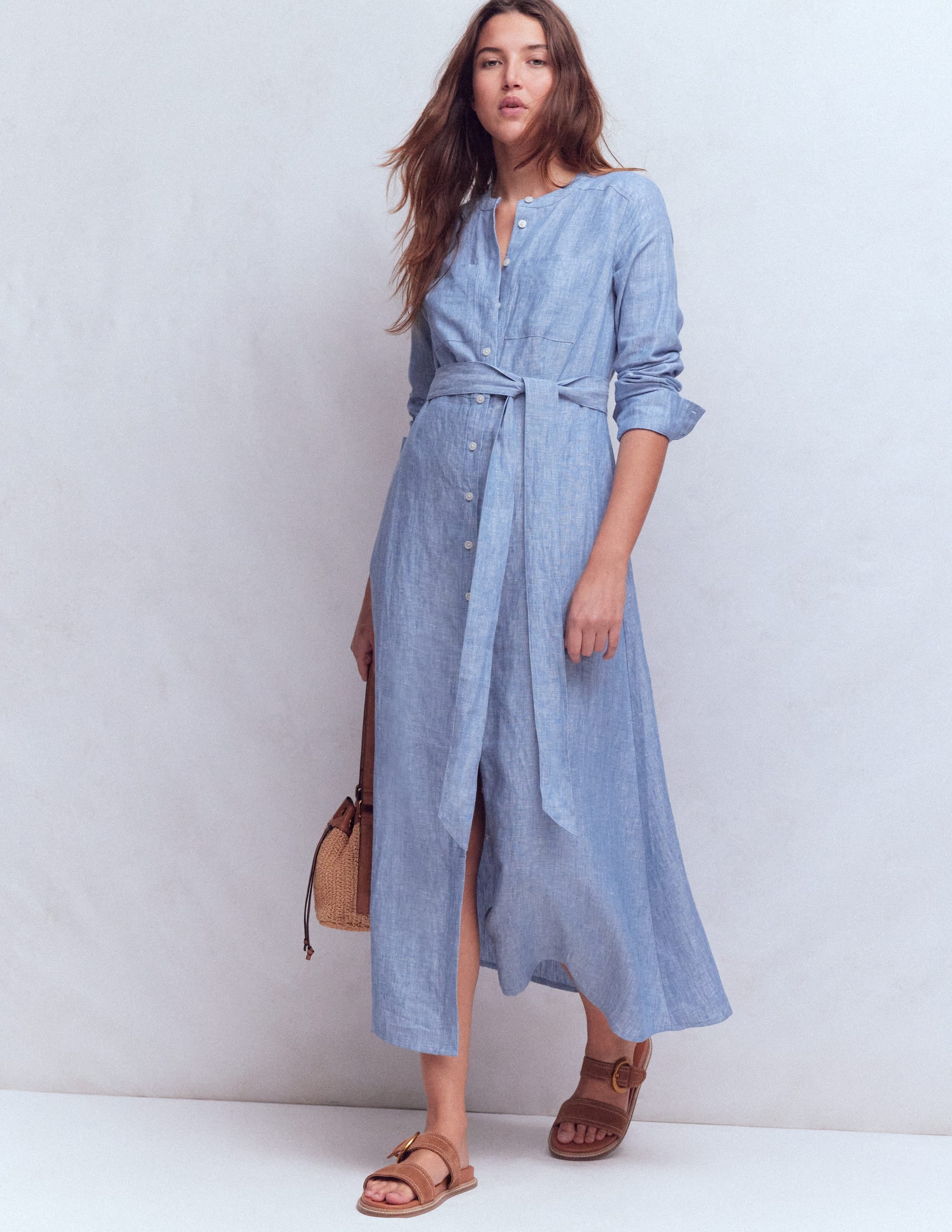 A woman wearing a light blue chambray linen shirt dress with a waist tie, long sleeves, and side pockets, paired with sandals and a woven handbag.